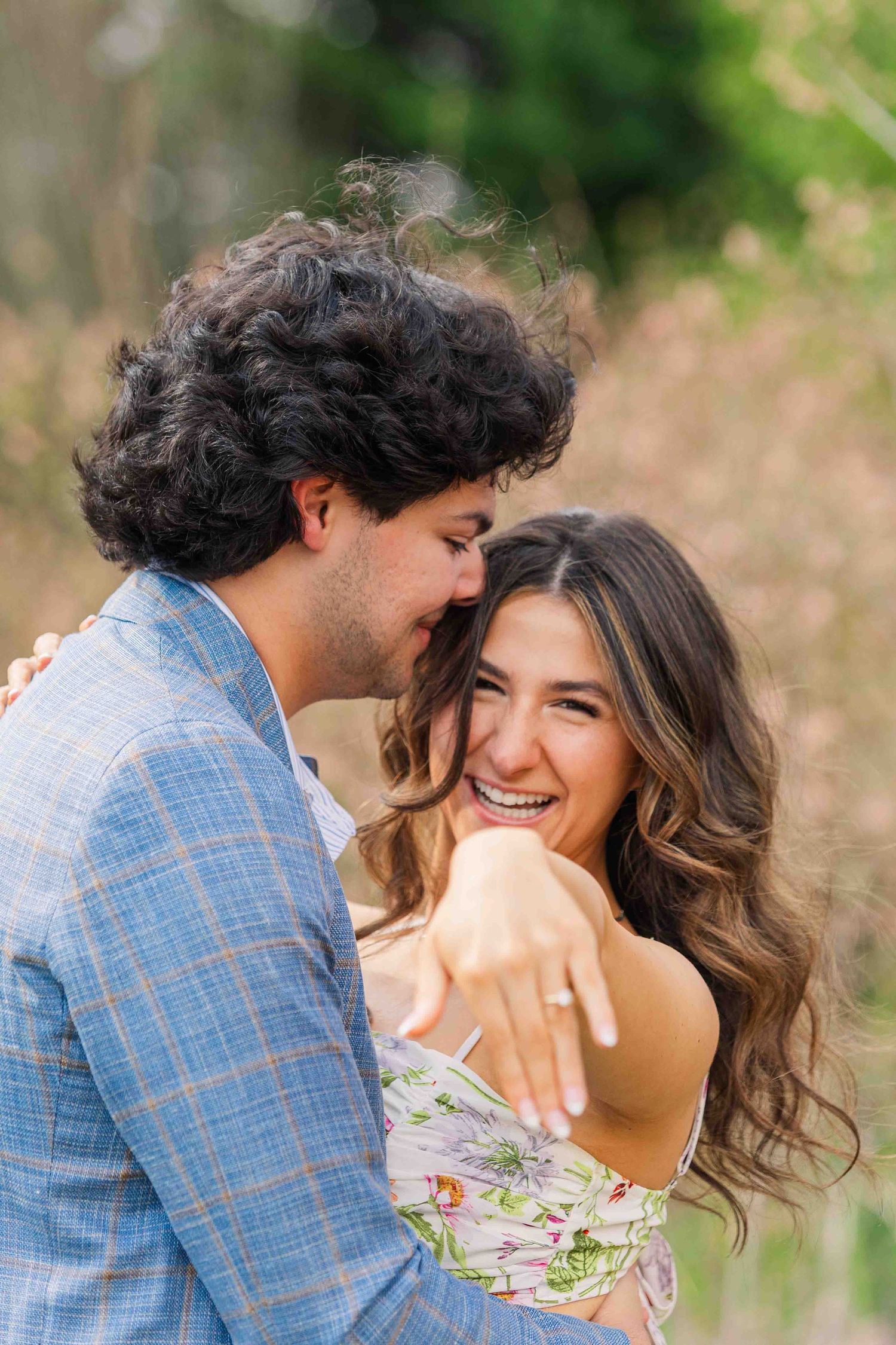 Smiling bride-to-be shows off her engagement ring while embracing her fiance in a sunlit outdoor setting.