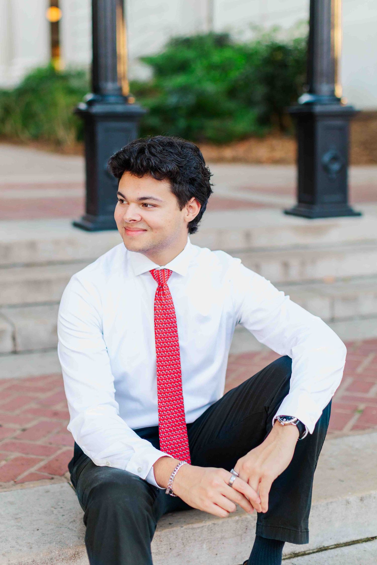 Young man in white dress shirt and red tie sits relaxed on outdoor steps smiling.