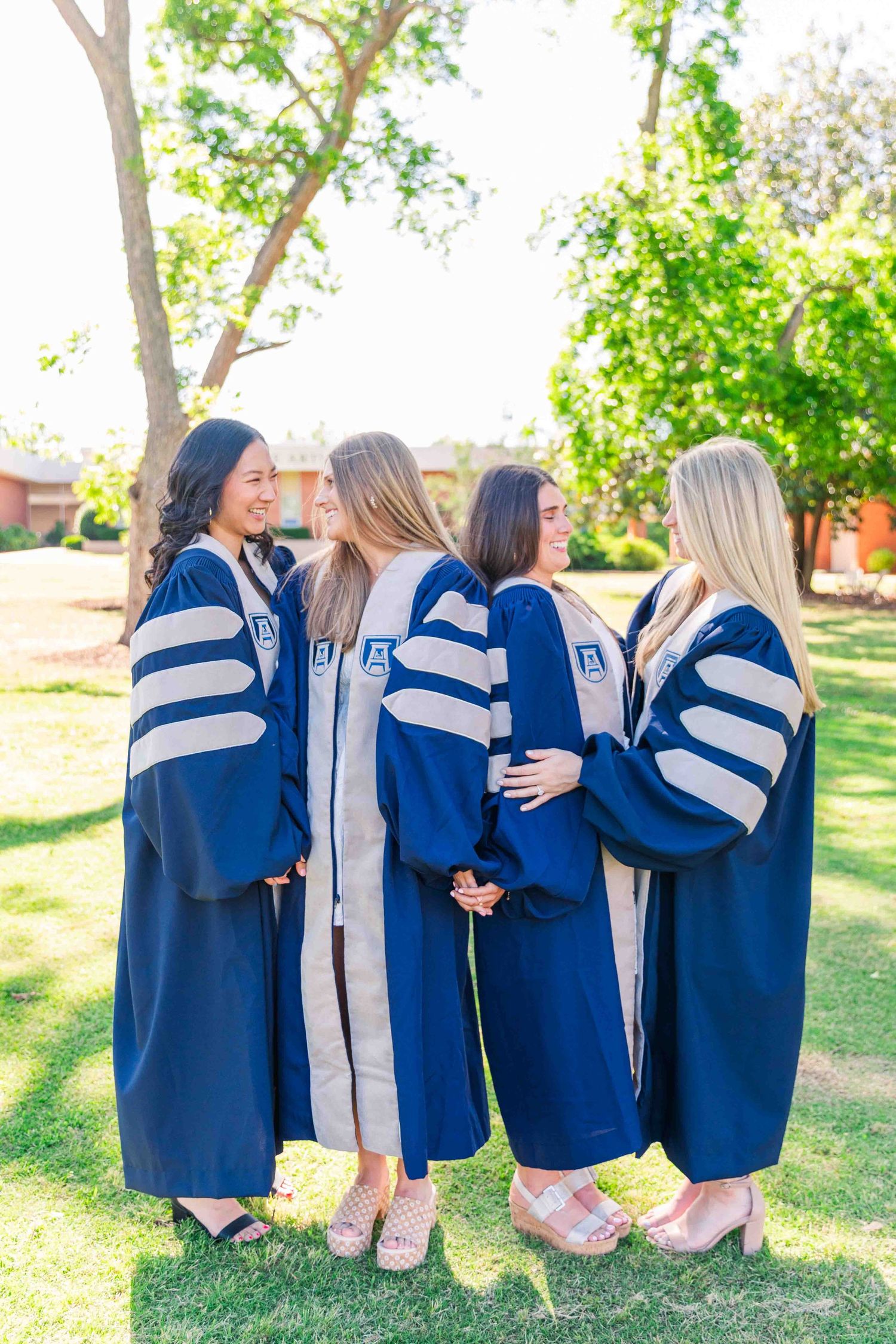 Four doctoral graduates in navy blue robes embrace from behind on green campus lawn.