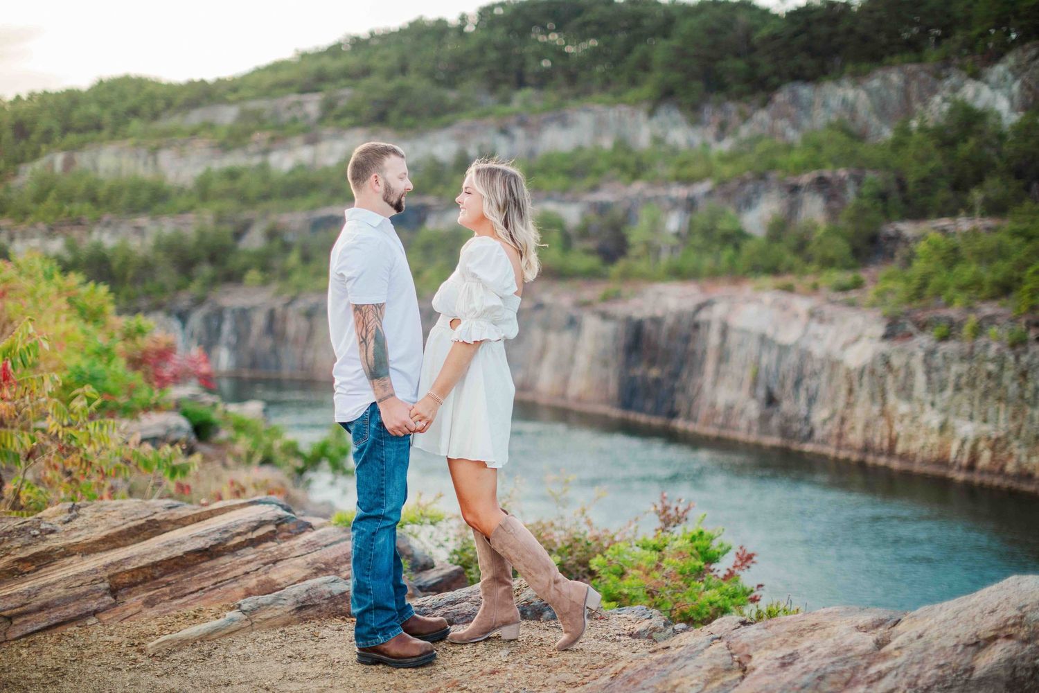 Couple holding hands on rocky cliff overlooking turquoise quarry water at sunset.