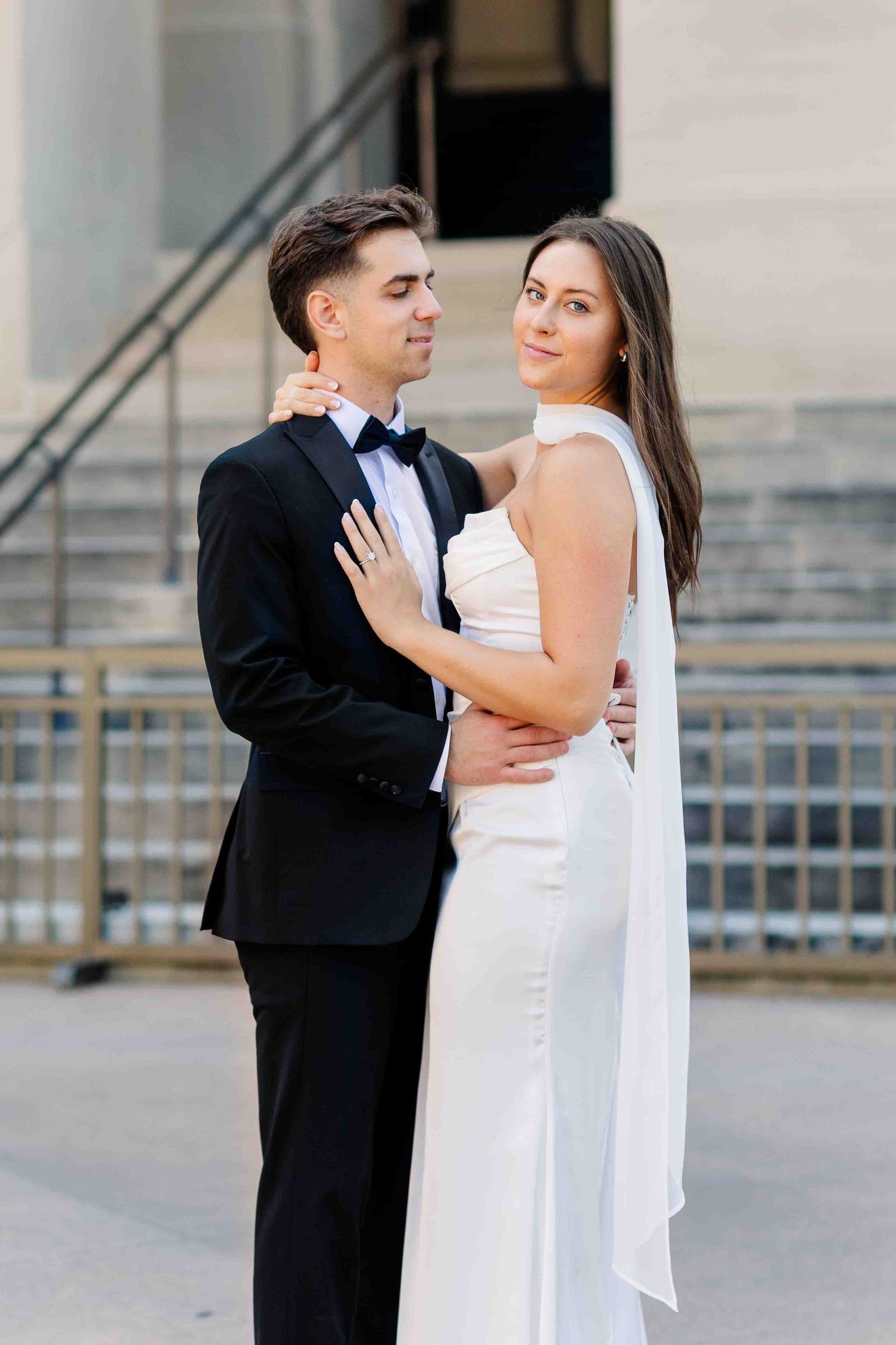 Elegant couple in wedding attire embrace outdoors, bride in white gown looks back, groom in black tuxedo.