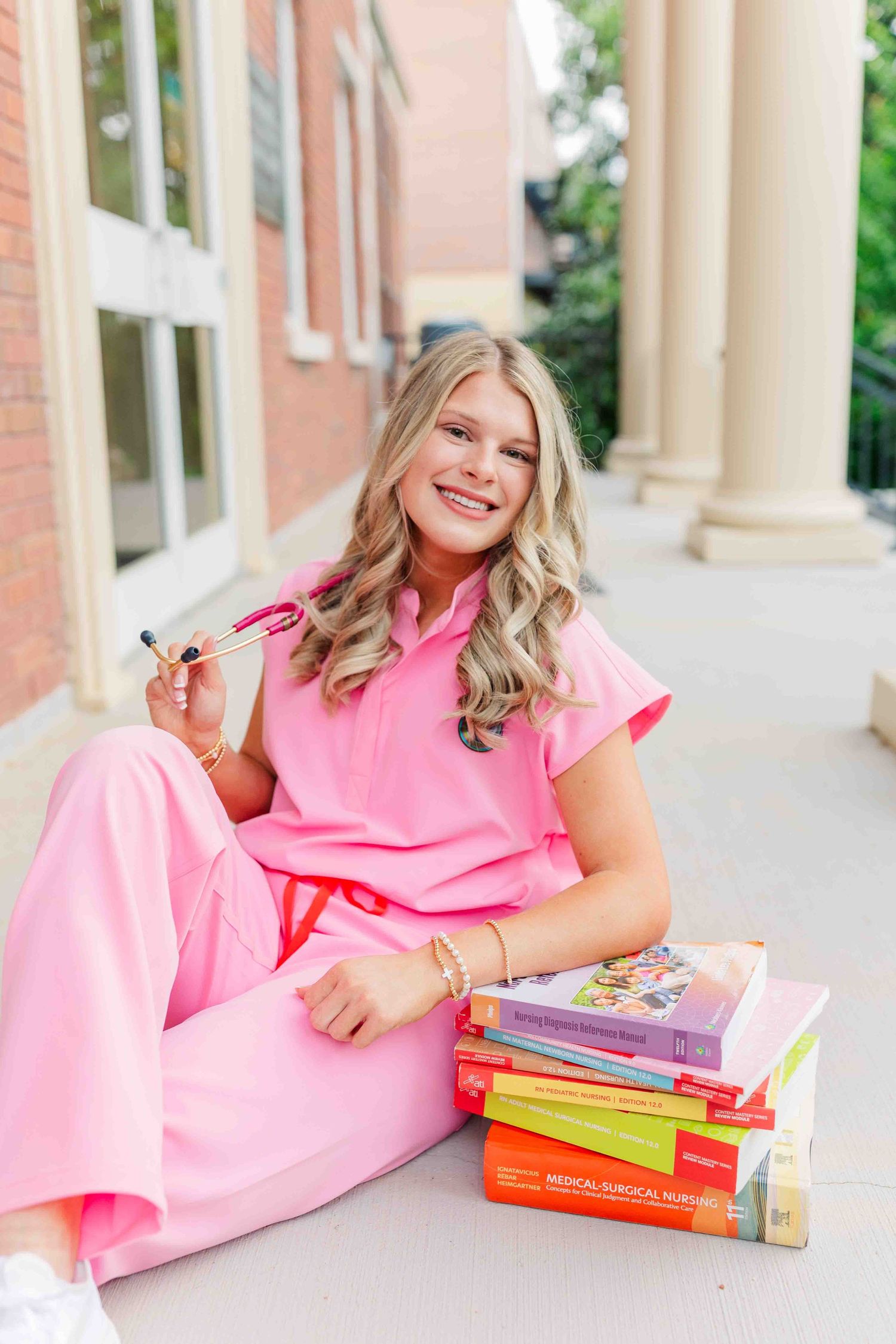 Smiling blonde nursing student in pink scrubs sitting with stacked medical textbooks on campus.