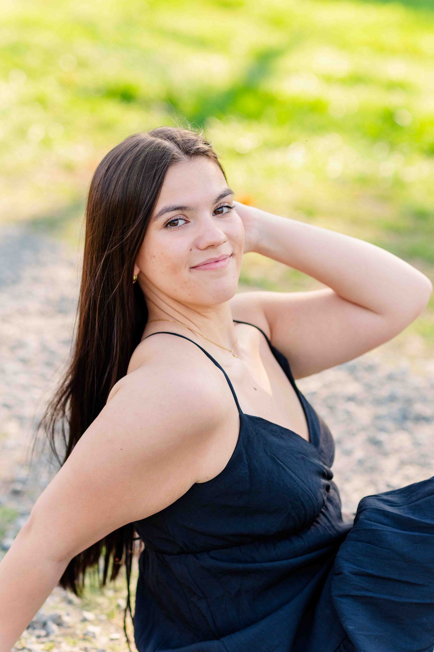 Smiling brunette woman in black dress sitting outdoors on sunny green grass path.