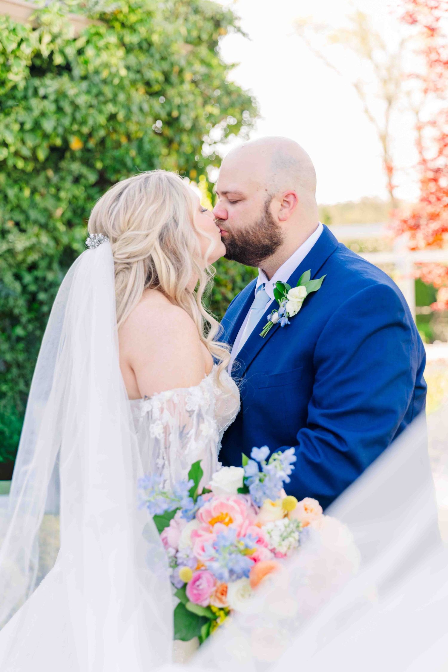 Bride and groom sharing a romantic kiss outdoors, bride holding a colorful bouquet with pink, blue, and yellow flowers.