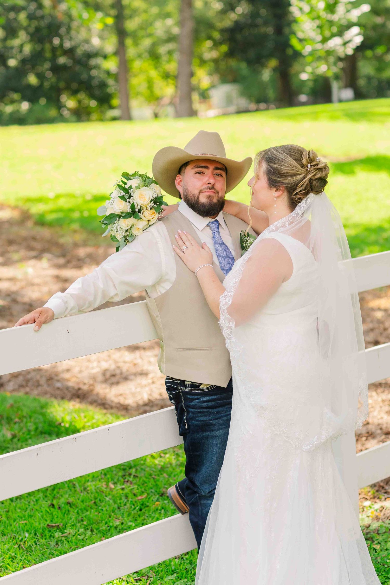 Bride in lace gown and groom in cowboy hat and vest pose by white fence at outdoor wedding.