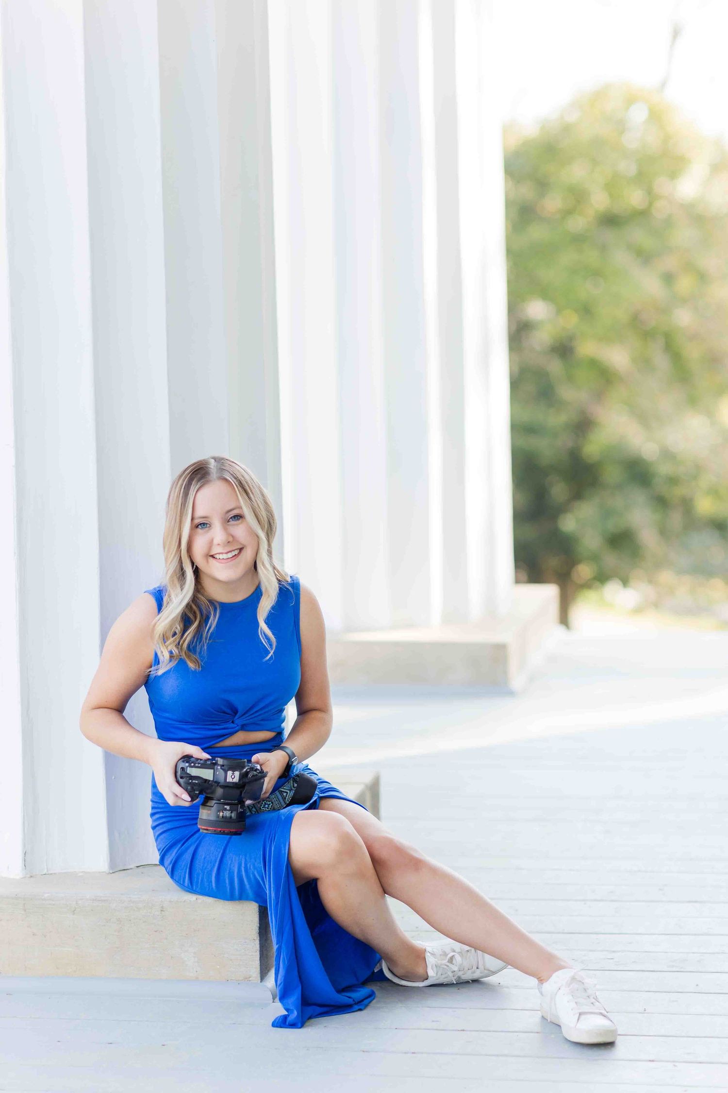 Professional female photographer in blue dress sitting relaxed with Canon DSLR camera near white pillars.