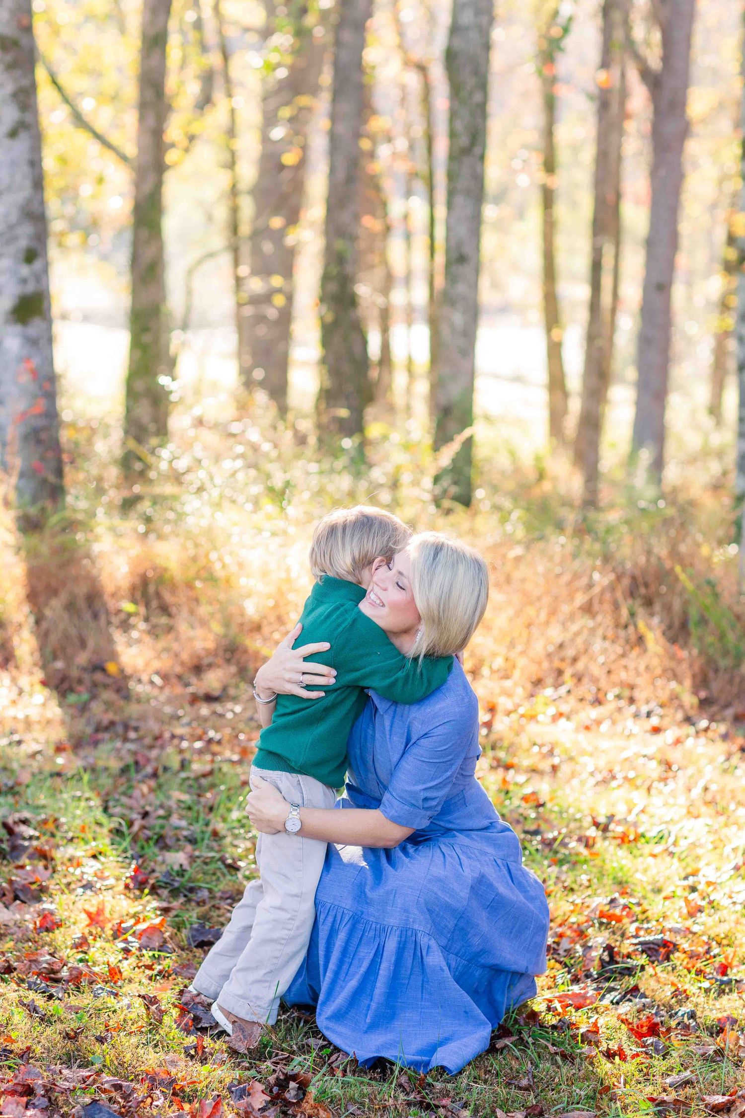 Blonde mother kneeling to embrace young son in sunny autumn forest with colorful leaves.