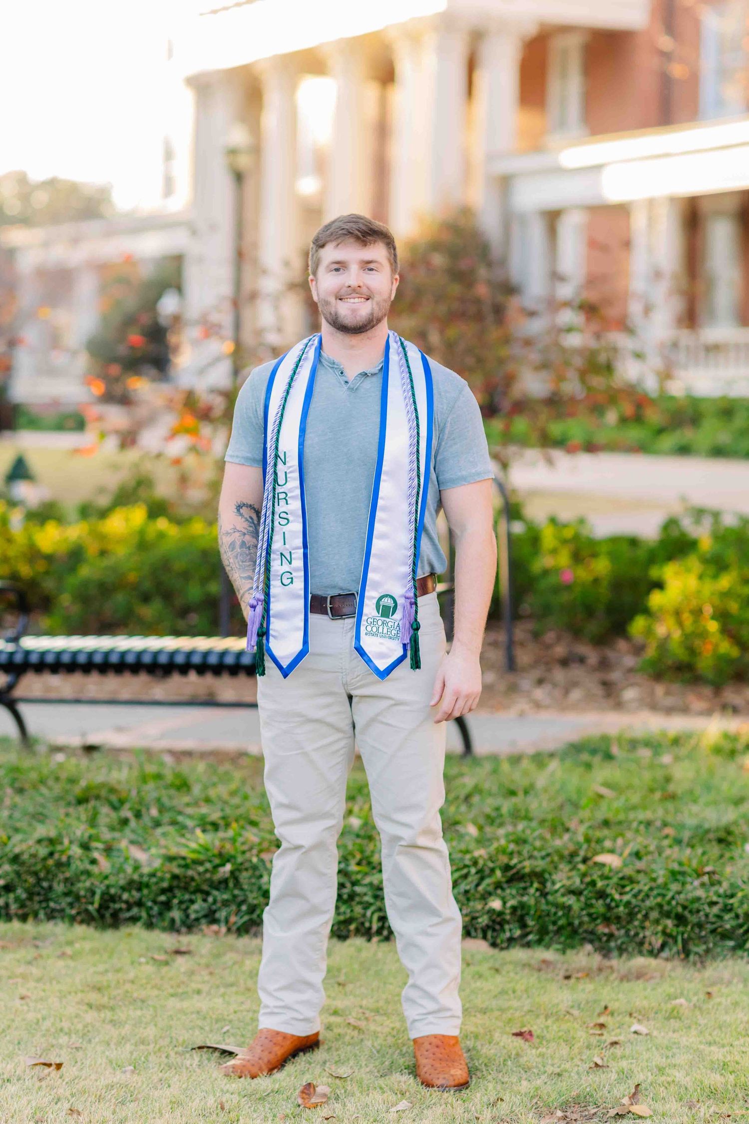 Smiling male graduate stands outdoors wearing blue stole and nursing graduation sash on campus.
