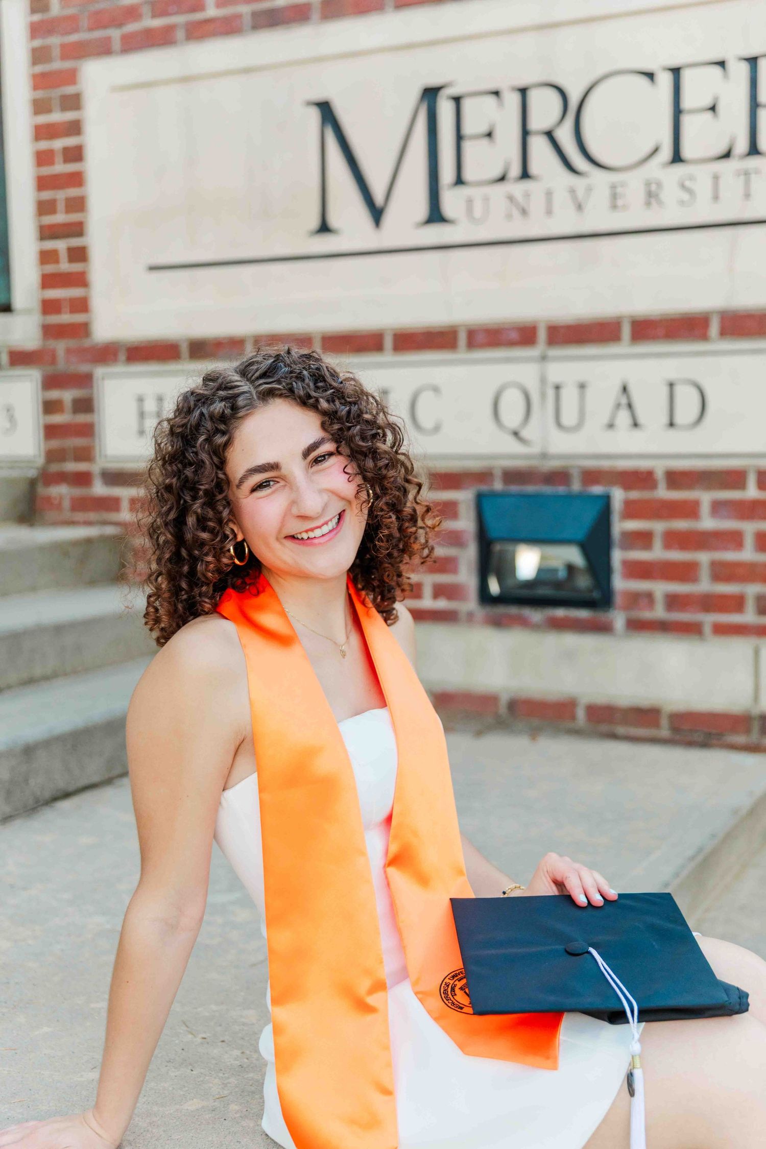 Smiling woman with curly hair in white dress wearing orange graduation stole at Mercer University.