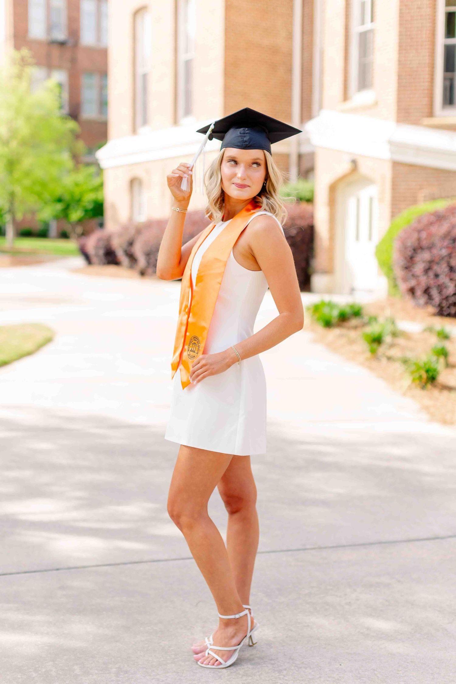 Blonde graduate in white mini dress, orange stole, and black cap posing on sunny campus path.