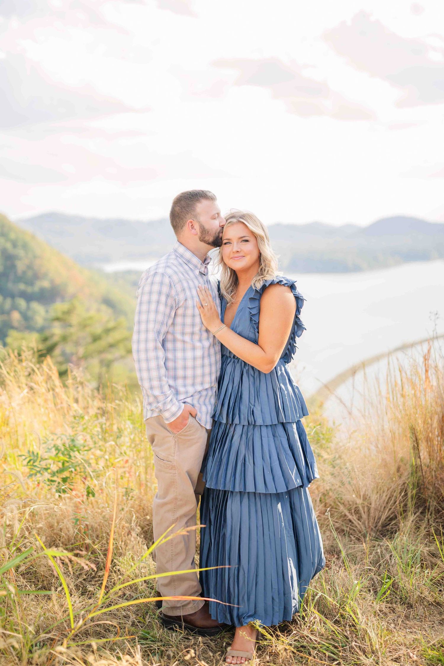 Couple posing on hillside overlook, man whispering to woman in blue tiered maxi dress at sunset.