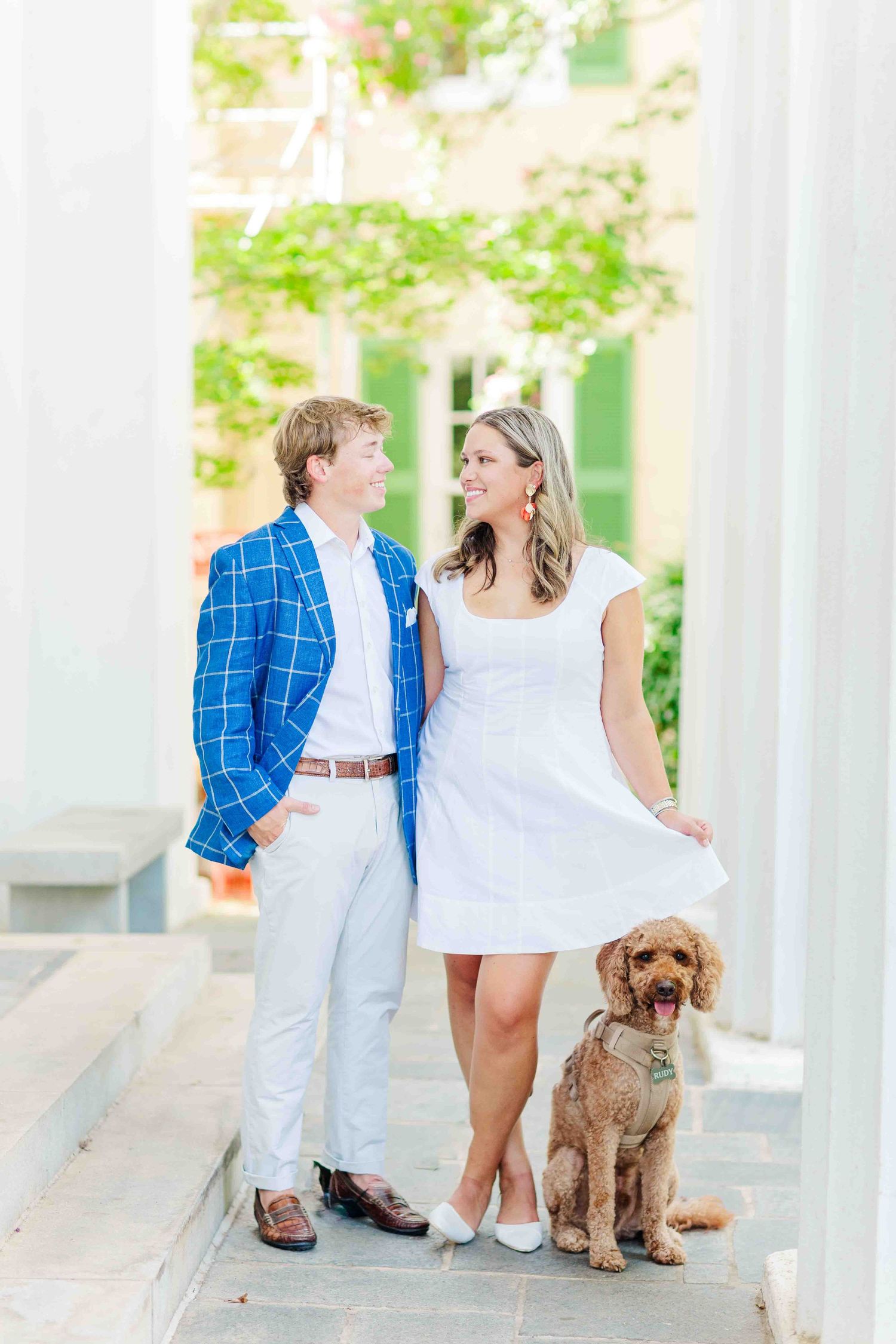 Smiling couple with their golden doodle dog posing between white columns in an outdoor setting.
