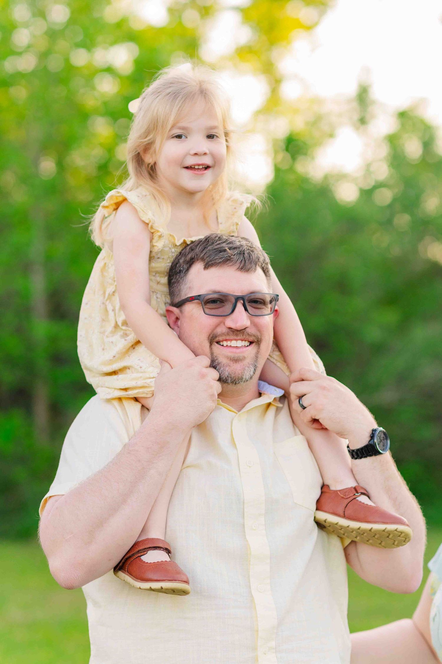 Smiling father wearing glasses carries his happy young blonde daughter on his shoulders outdoors.