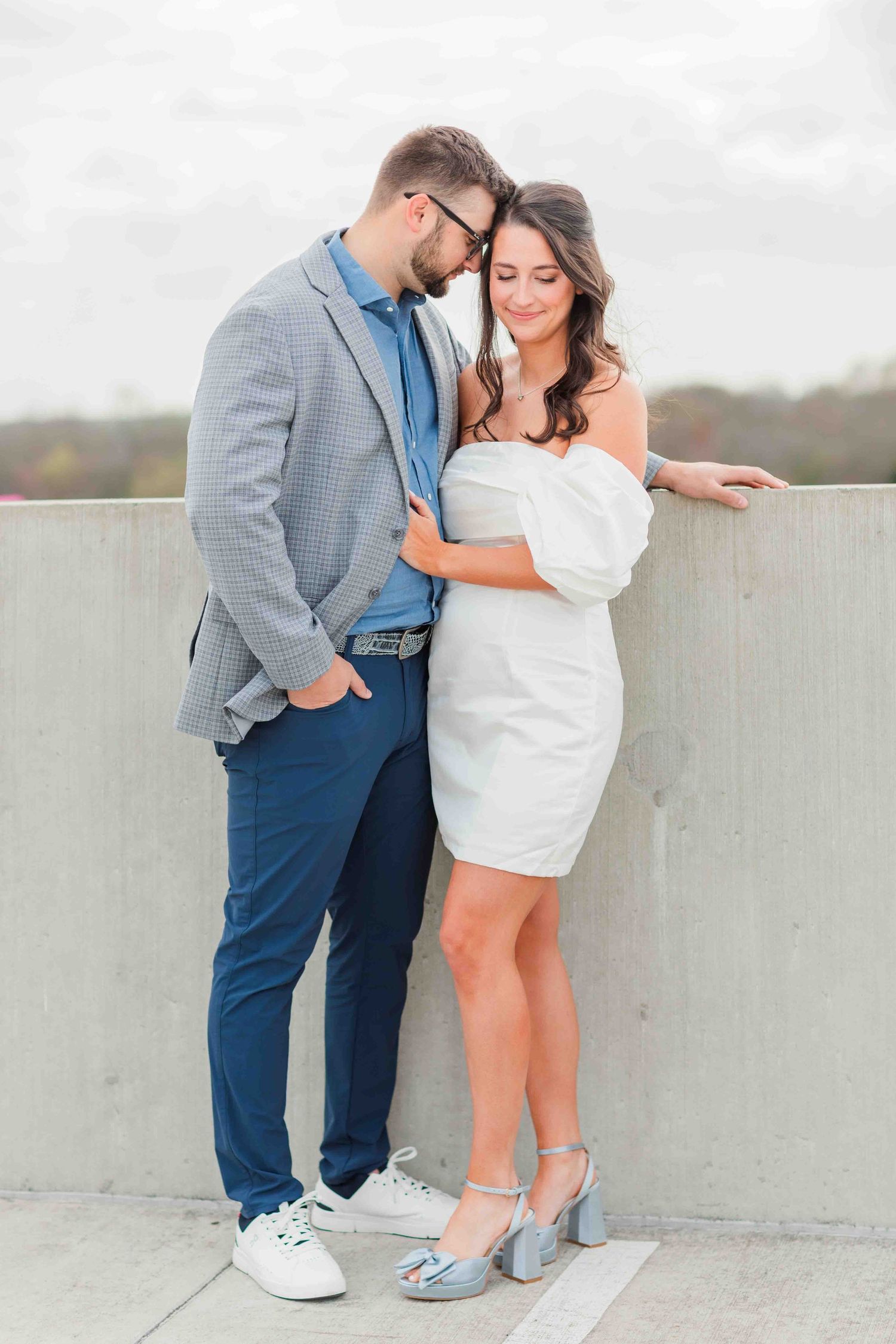 Engaged couple posing on rooftop, man in gray blazer and woman in white off-shoulder mini dress.
