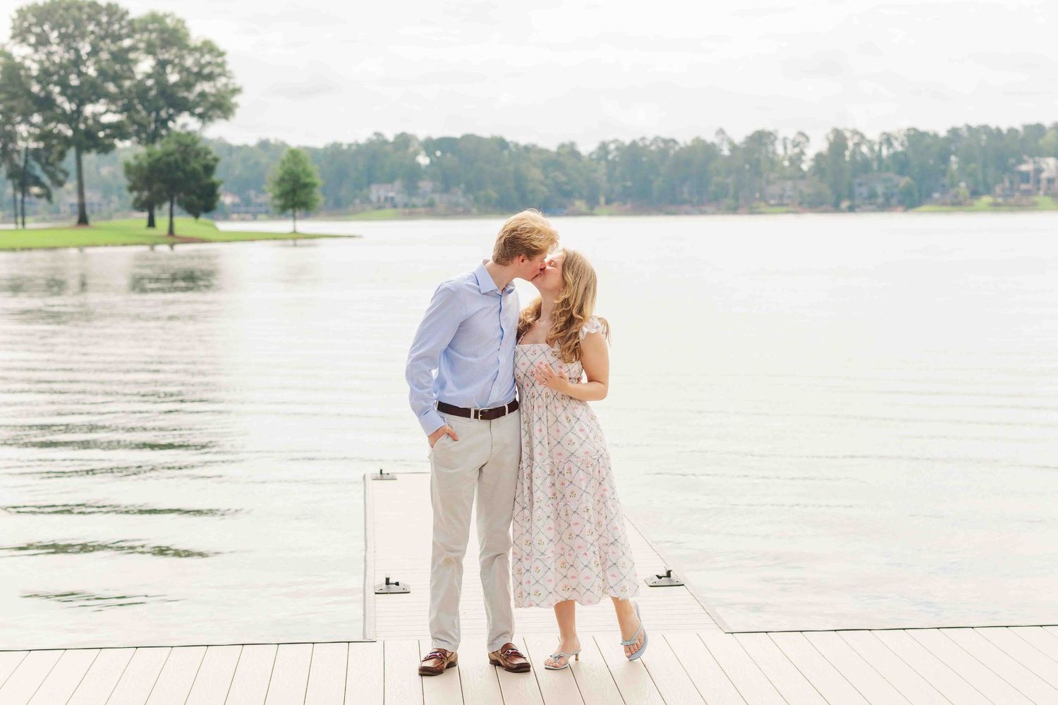 Couple sharing a kiss on a wooden dock overlooking a serene lake surrounded by lush green trees.