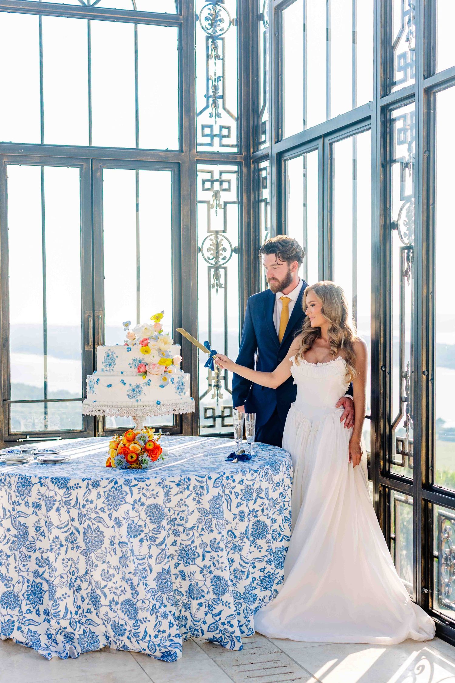 Bride and groom cutting a blue and white floral wedding cake at an elegant venue with large ornate windows.