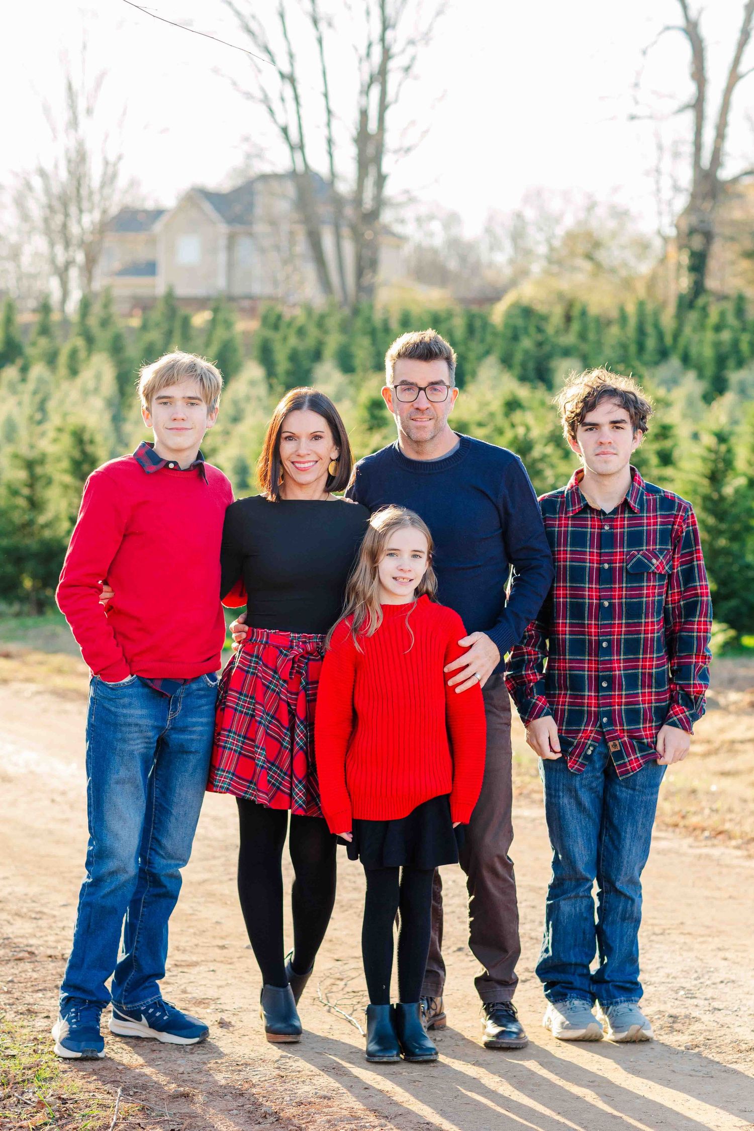 Family of five in red and navy outfits posing together at a Christmas tree farm on a sunny day.