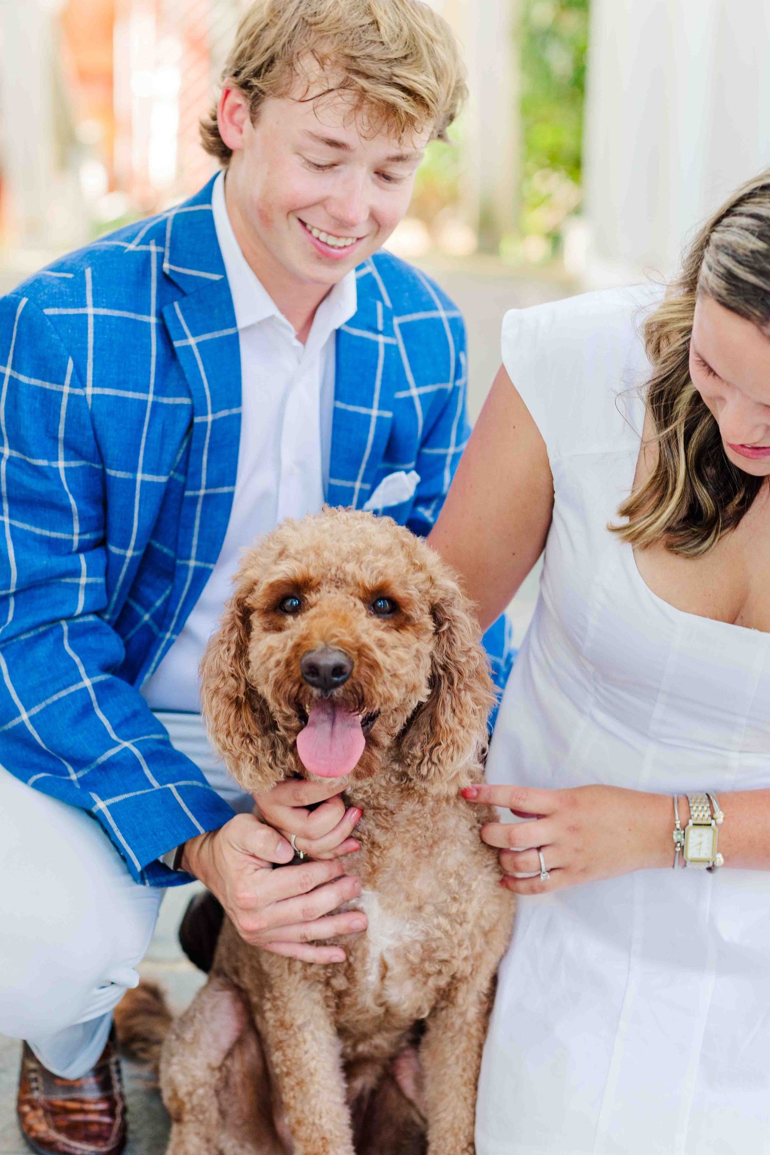Smiling couple in blue plaid blazer and white dress posing with fluffy golden doodle dog outdoors.