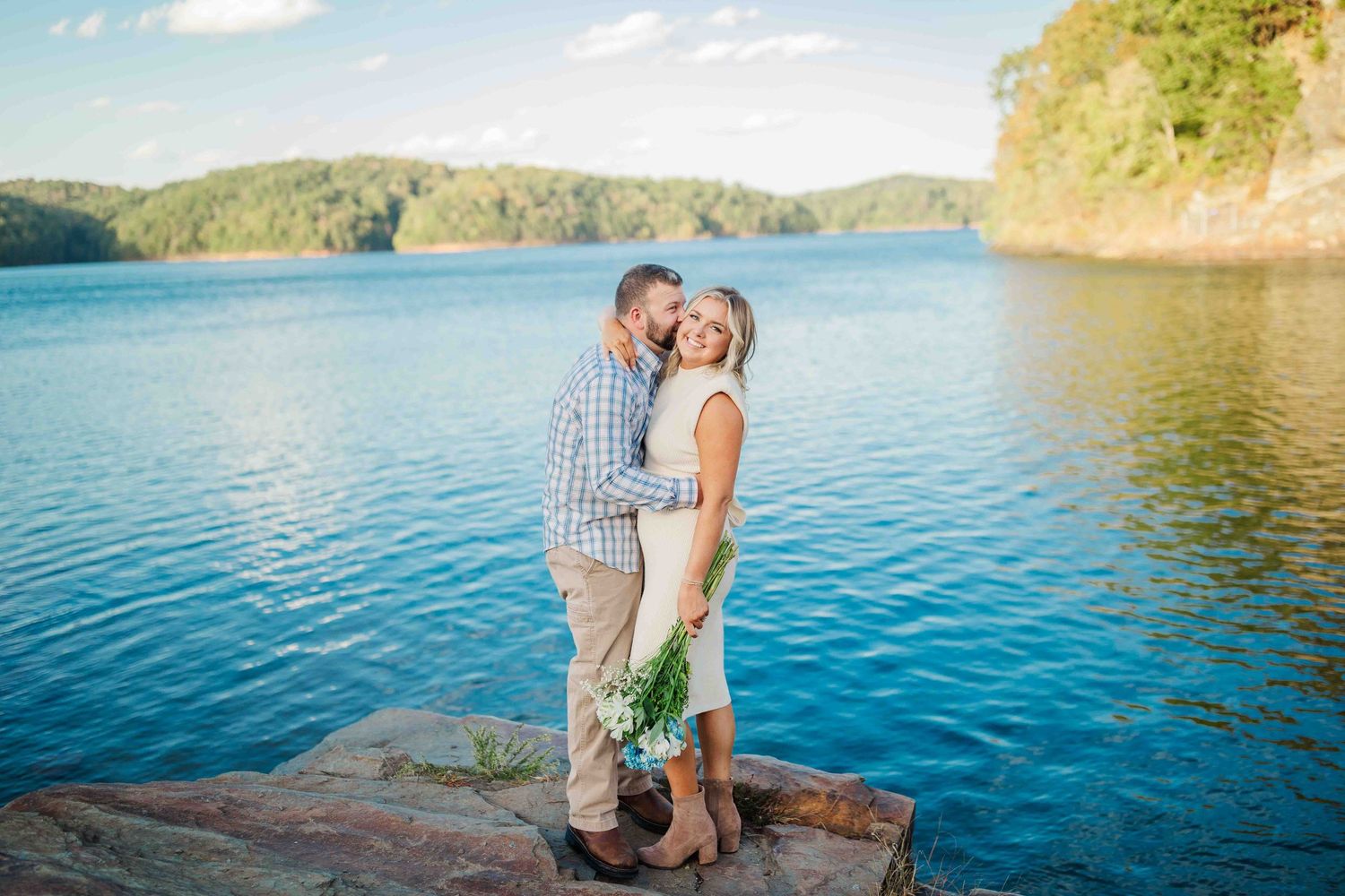Couple kissing on rocky lakeside, woman holding white wildflowers with vibrant blue water behind them.