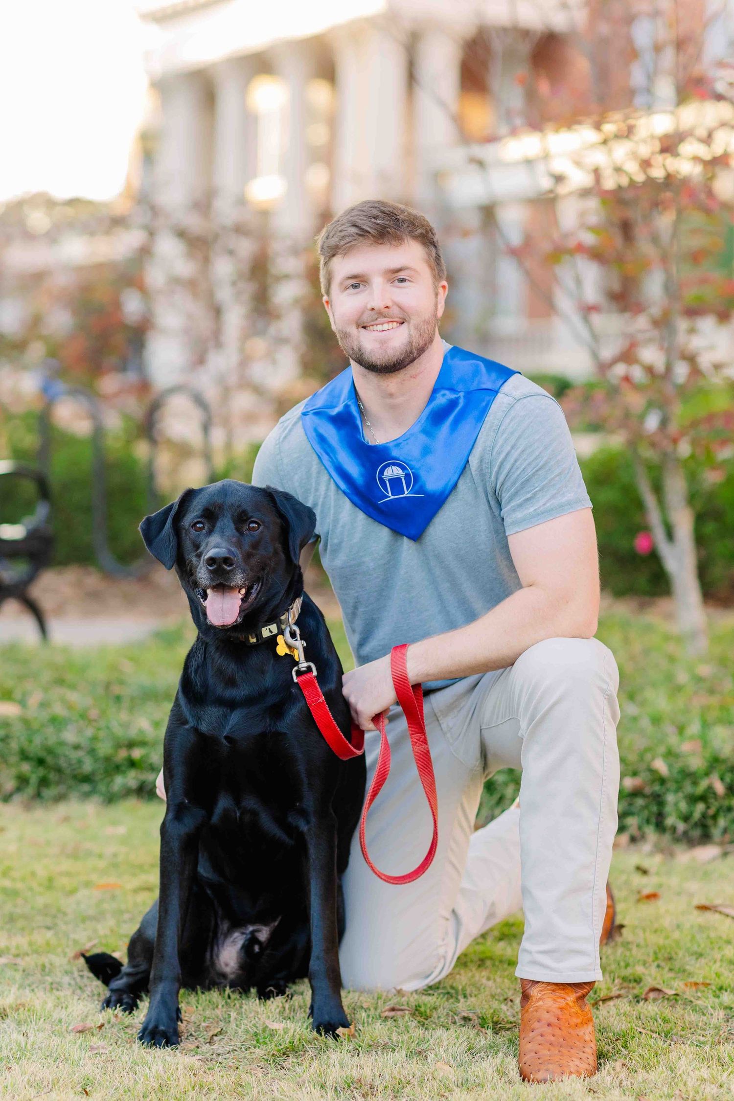 Smiling male graduate kneels beside black Labrador Retriever on red leash on campus lawn.