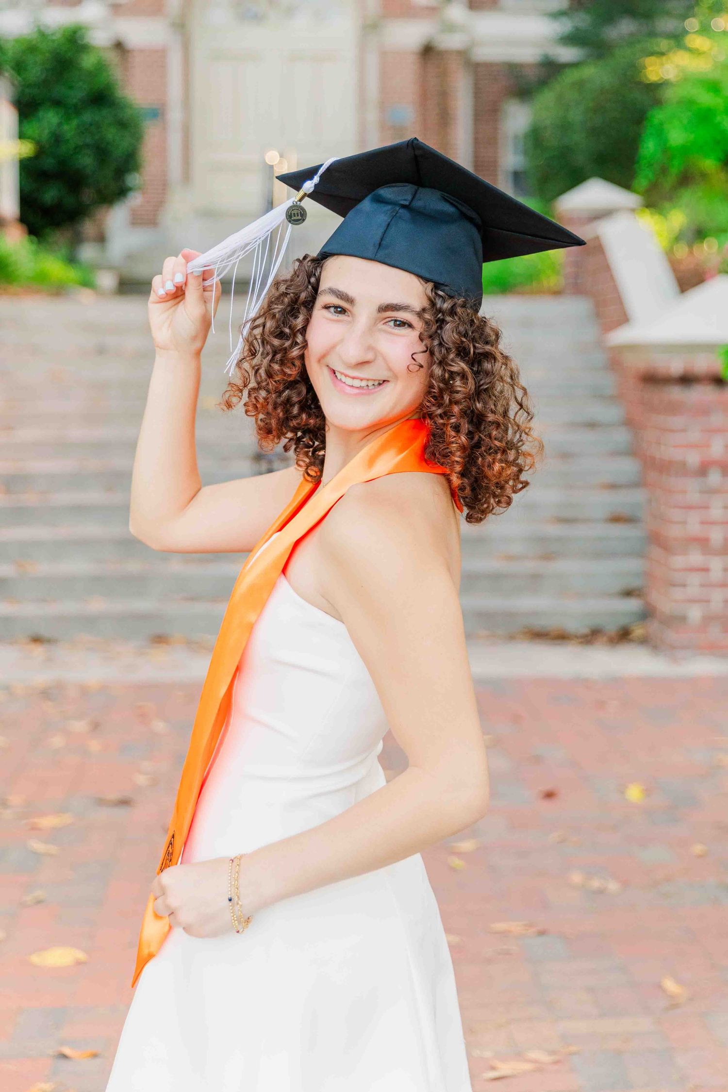 Smiling graduate in black cap and orange sash poses in white dress on brick campus steps.