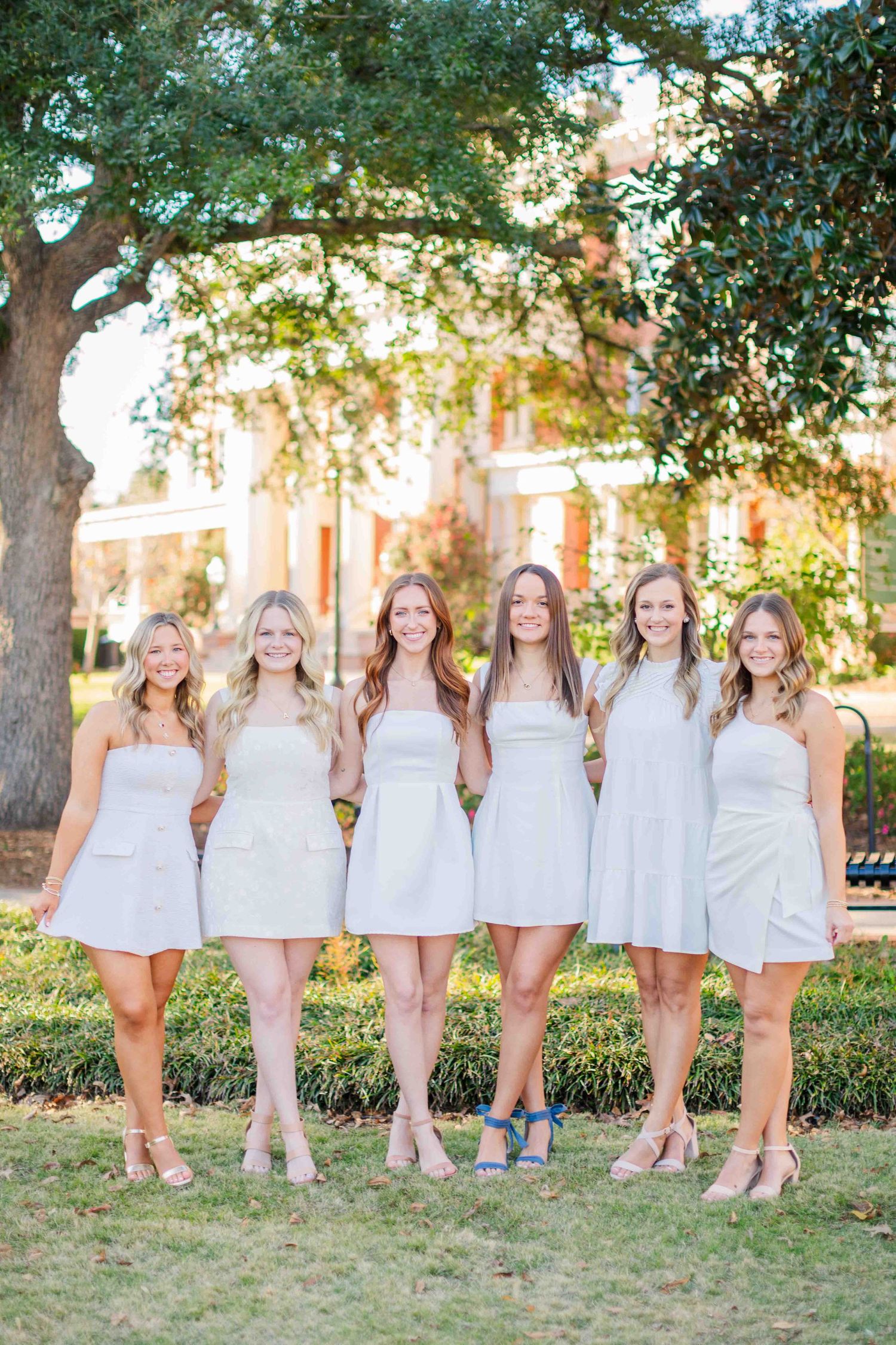 Six young women in matching white mini dresses pose together outdoors on a sunny day near a historic building.