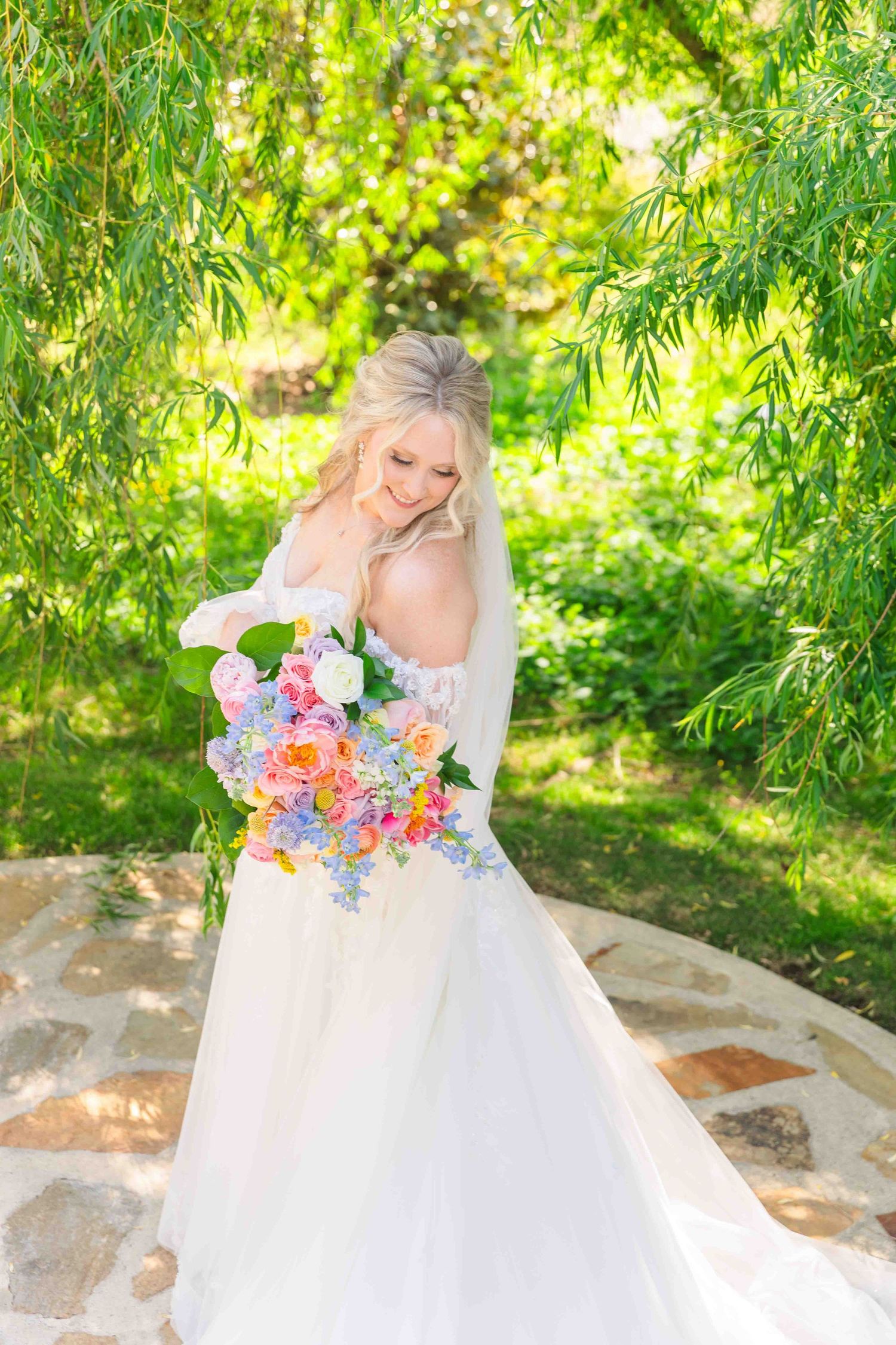 Blonde bride in white gown holds colorful bouquet under weeping willow tree in garden.