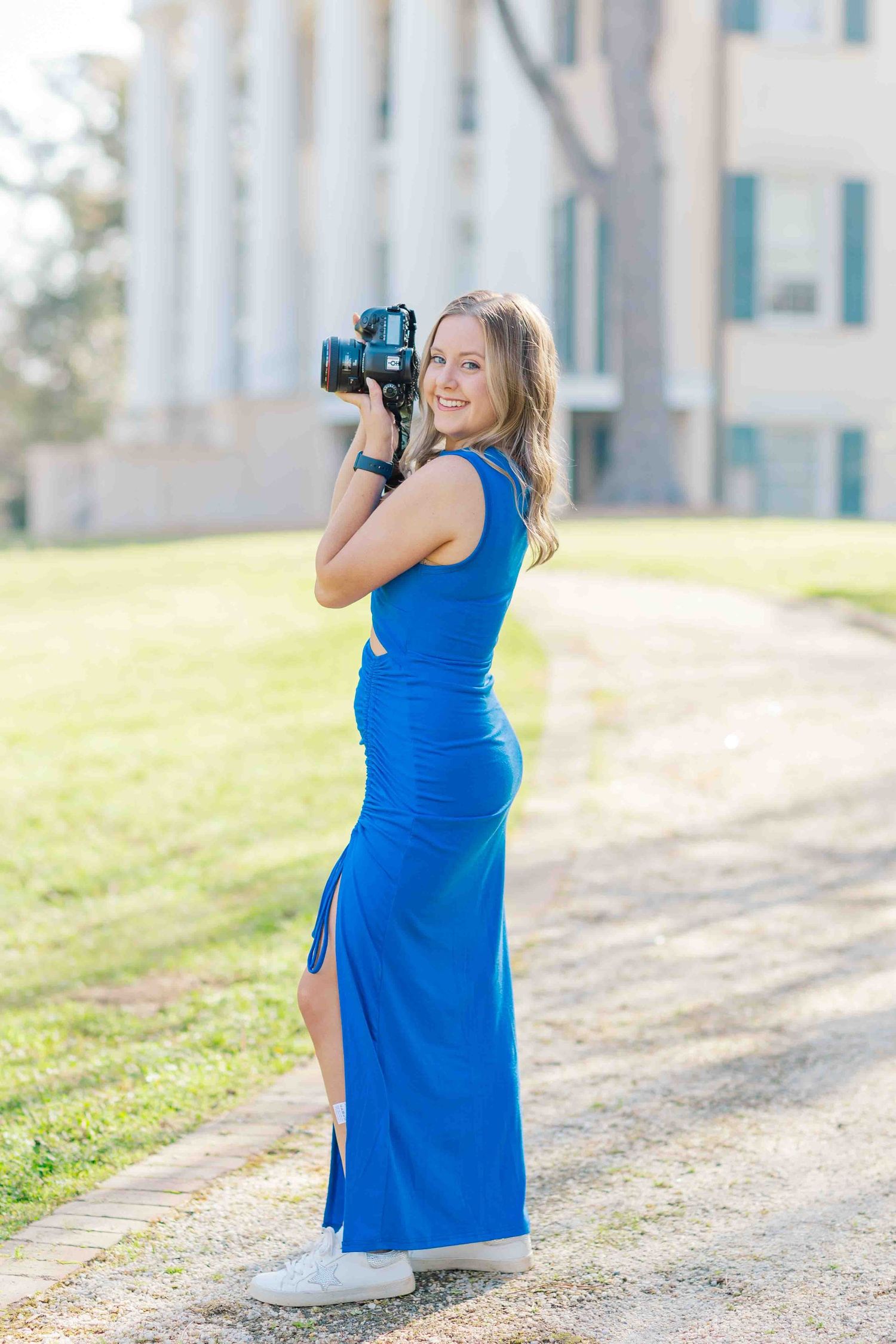Smiling young woman in blue dress holding DSLR camera on sunlit campus pathway.