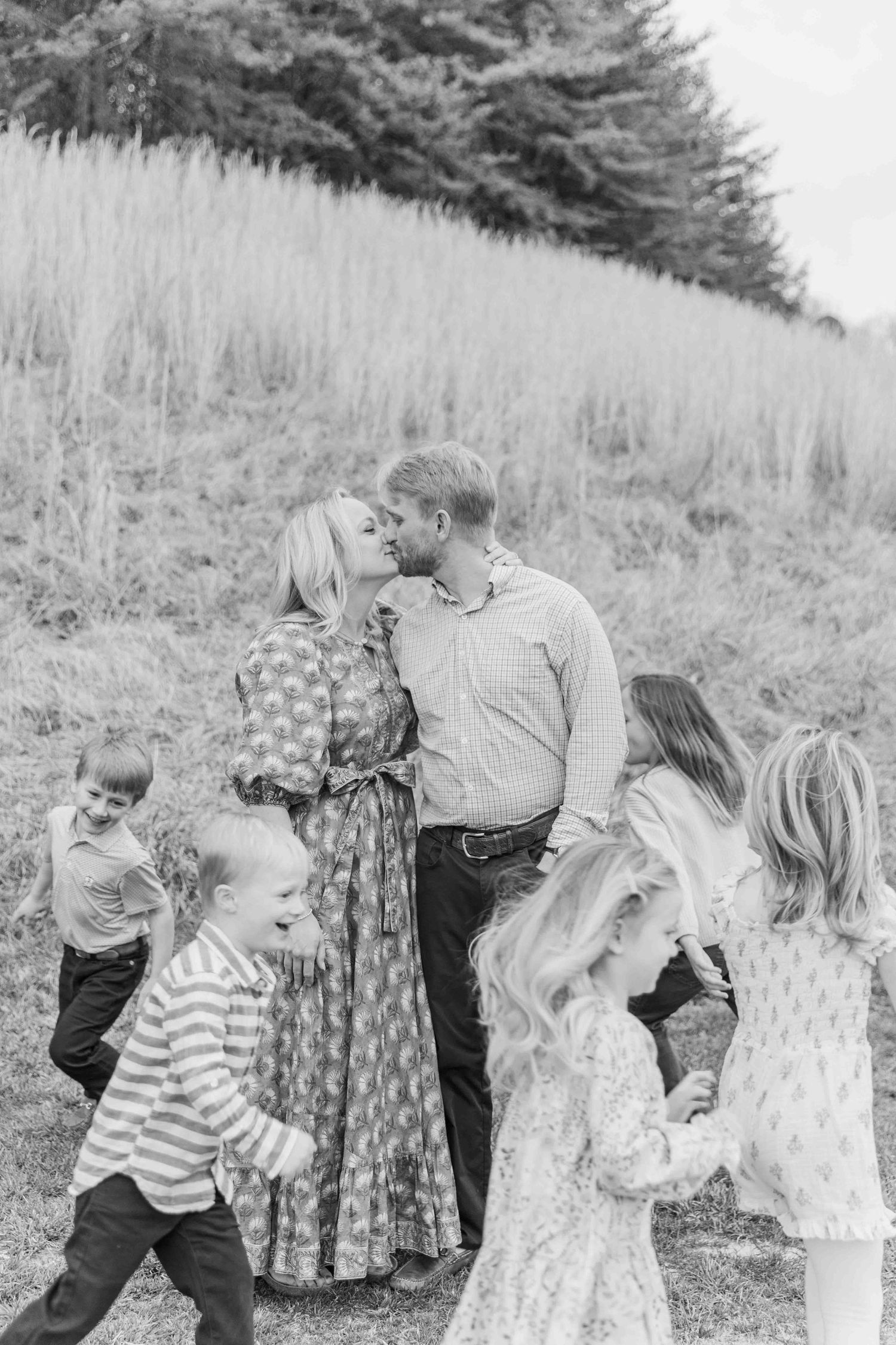 Black and white family photo of parents kissing surrounded by four young children in a meadow.