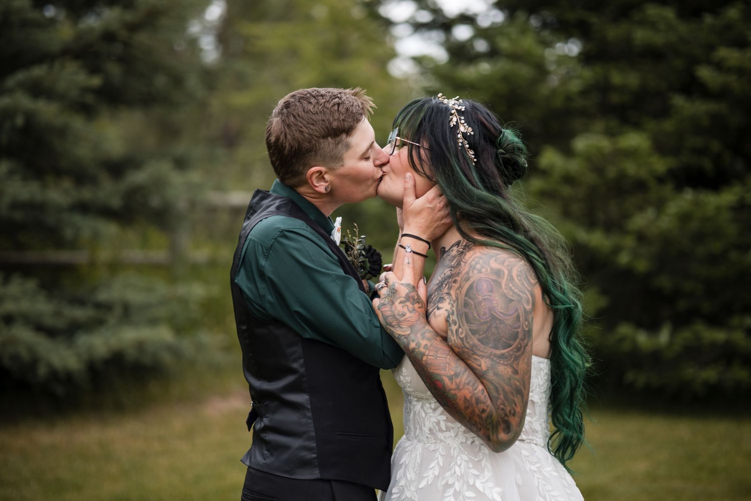 Tattooed bride and groom sharing first kiss outdoors with evergreen trees behind.