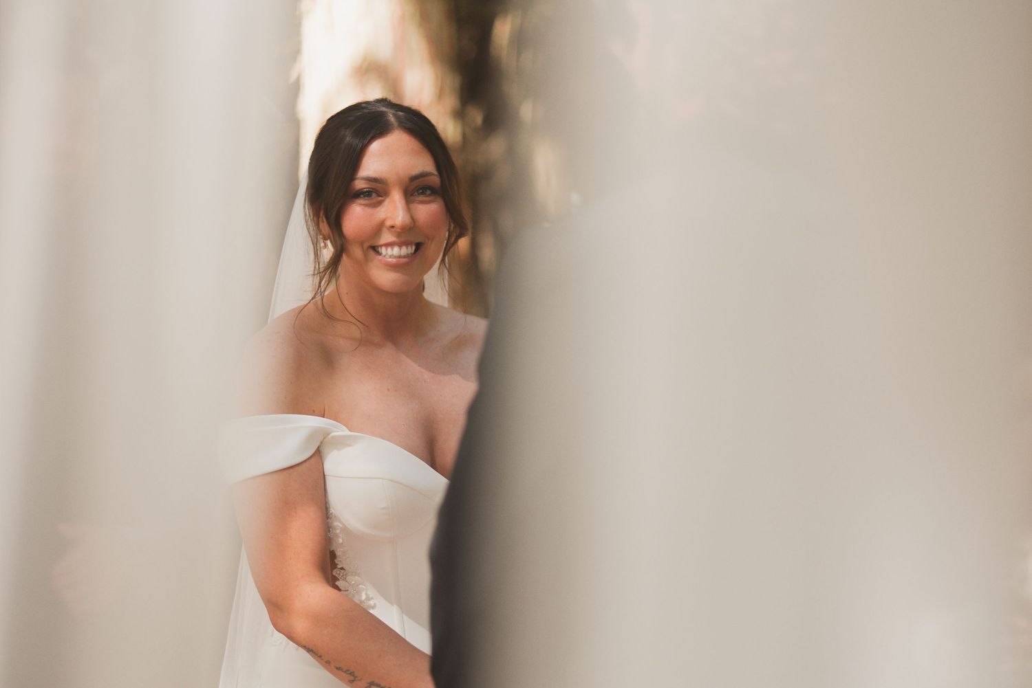 Smiling bride in an off-shoulder white gown and veil, peeking between white columns.