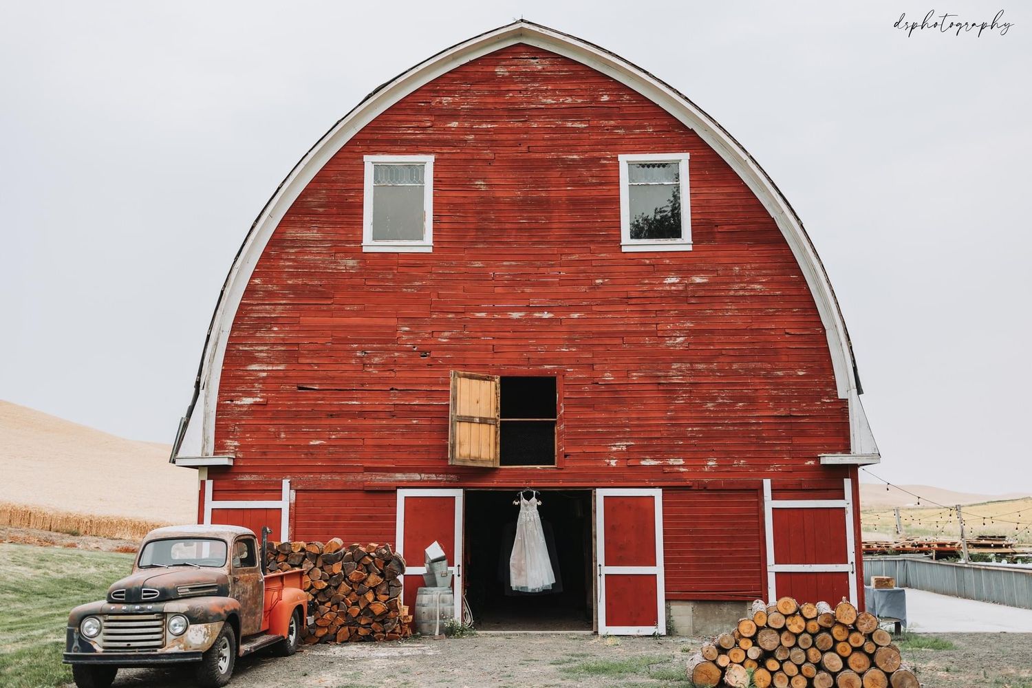 White wedding dress hanging in the doorway of a rustic red barn with stacked logs.