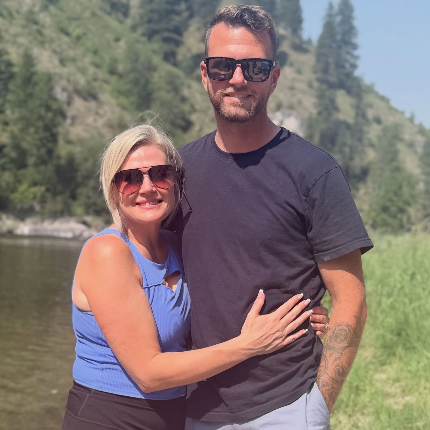 Couple posing together near a scenic river with lush green mountain hills behind them.