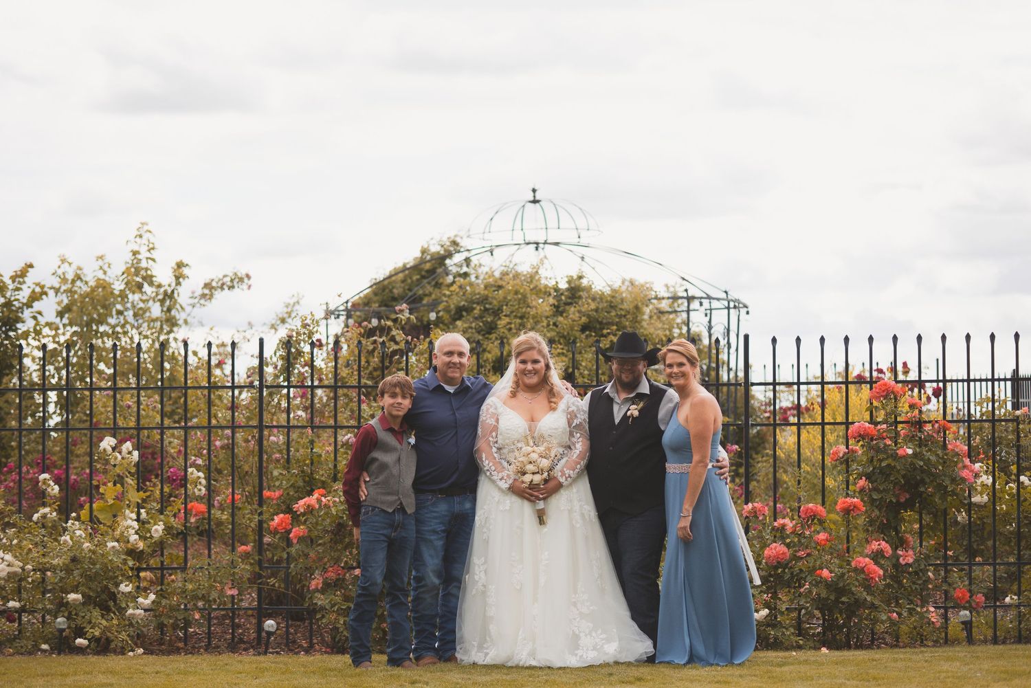 Wedding group photo of bride, groom, and guests posing before an iron fence with roses.