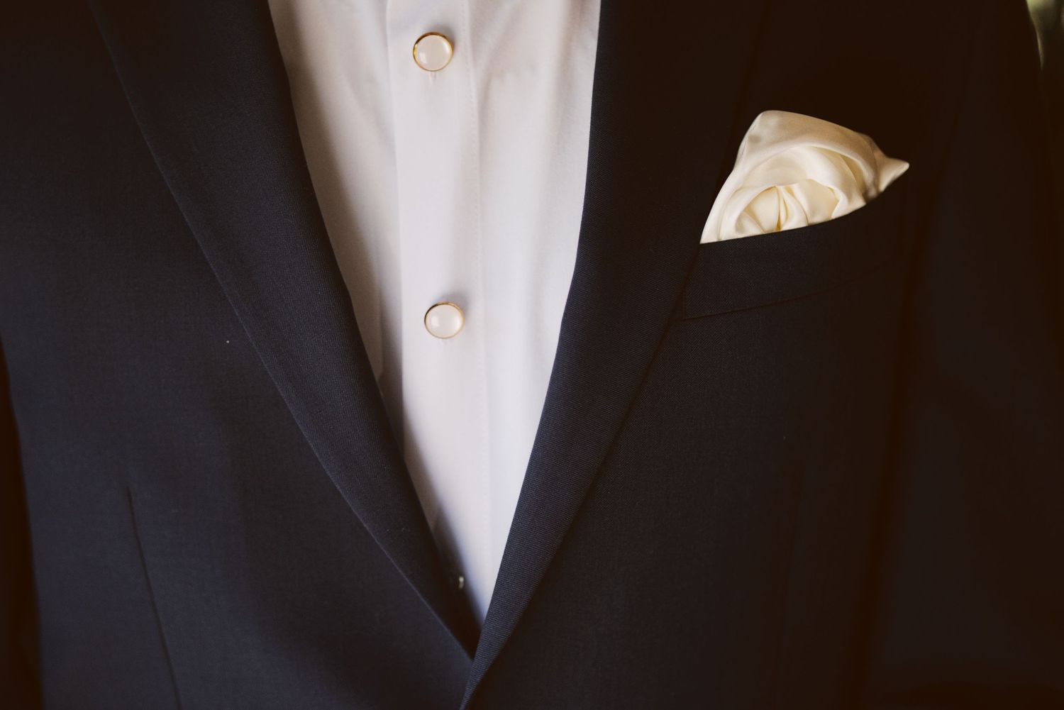 Close-up of a groom's black tuxedo with white shirt, gold buttons, and white pocket square.