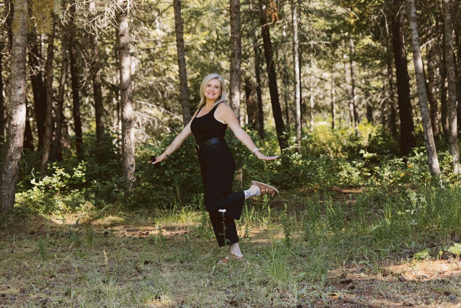 Pregnant woman in black outfit posing outdoors in a sunlit forest clearing.