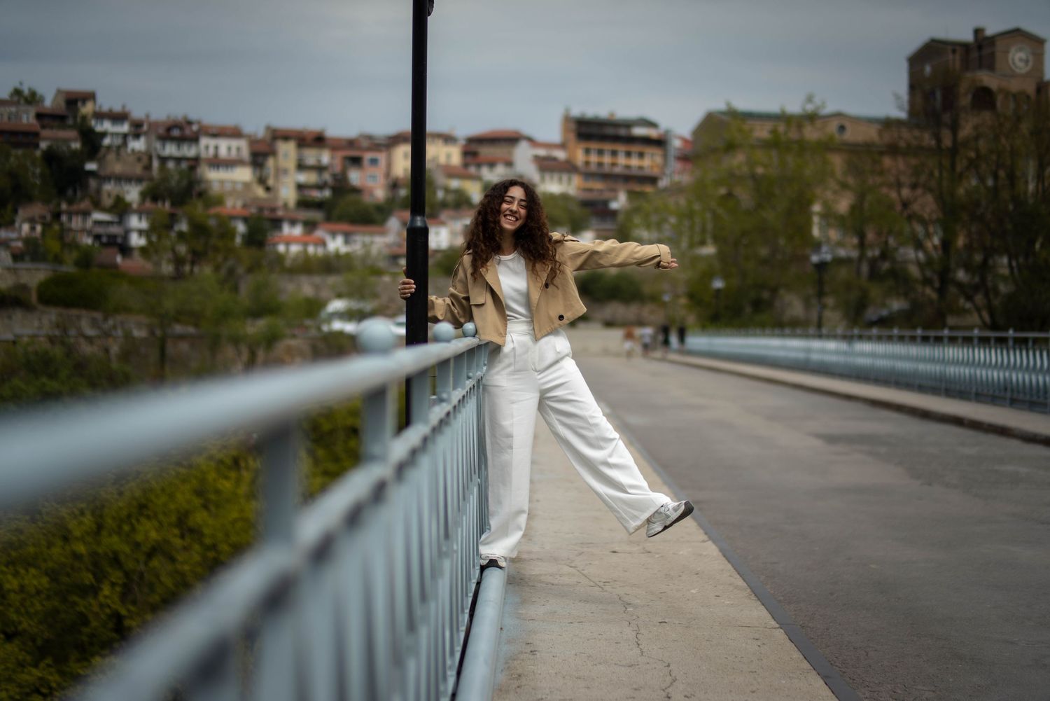 Woman in beige jacket and white pants posing on bridge with hillside town in background.