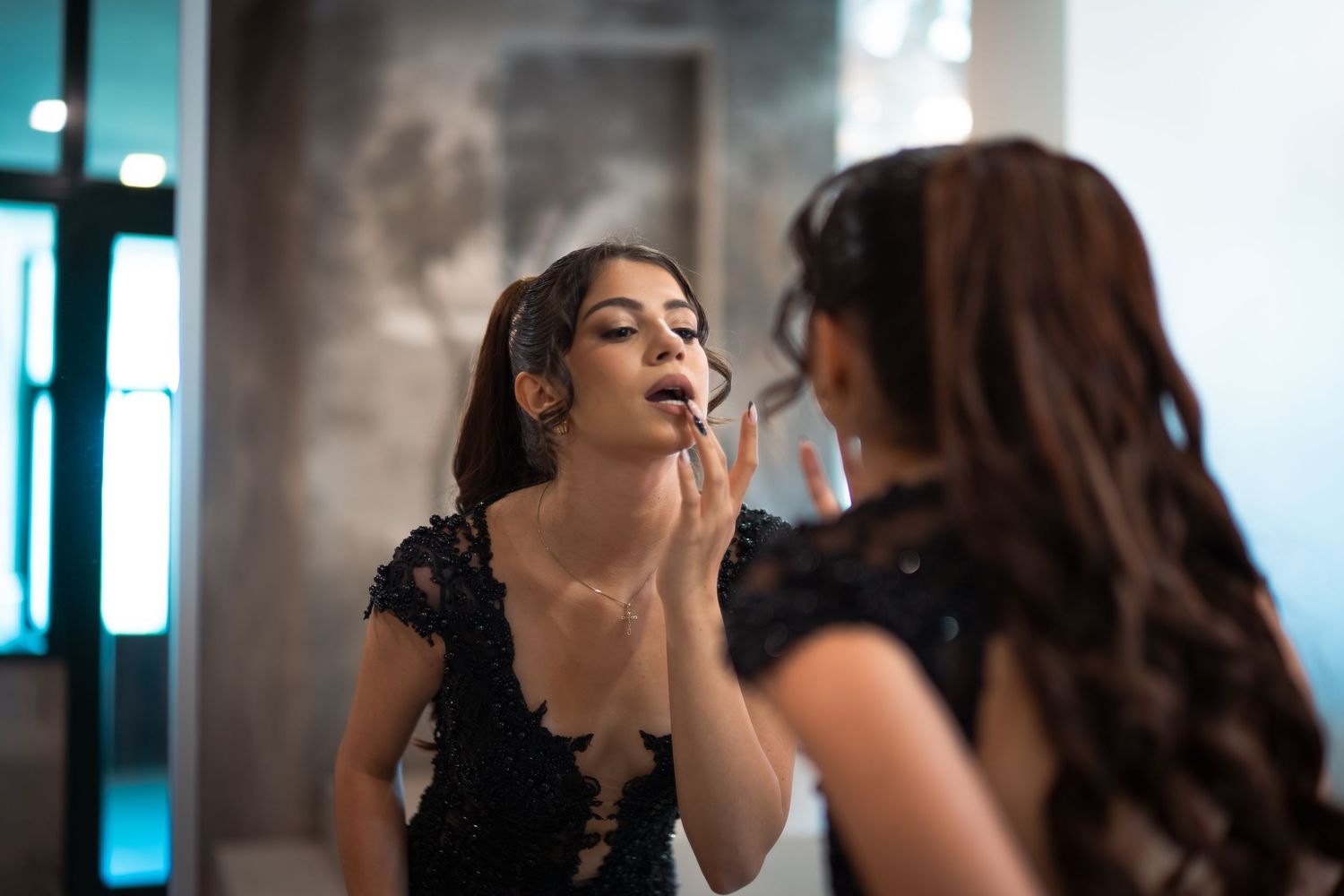 Young woman applying lipstick while looking in mirror, wearing black lace dress.