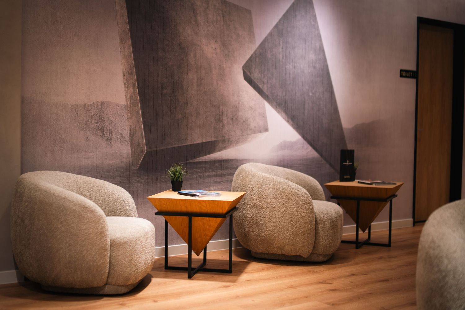 Modern waiting area with two rounded beige armchairs and wooden side tables against abstract wall mural.