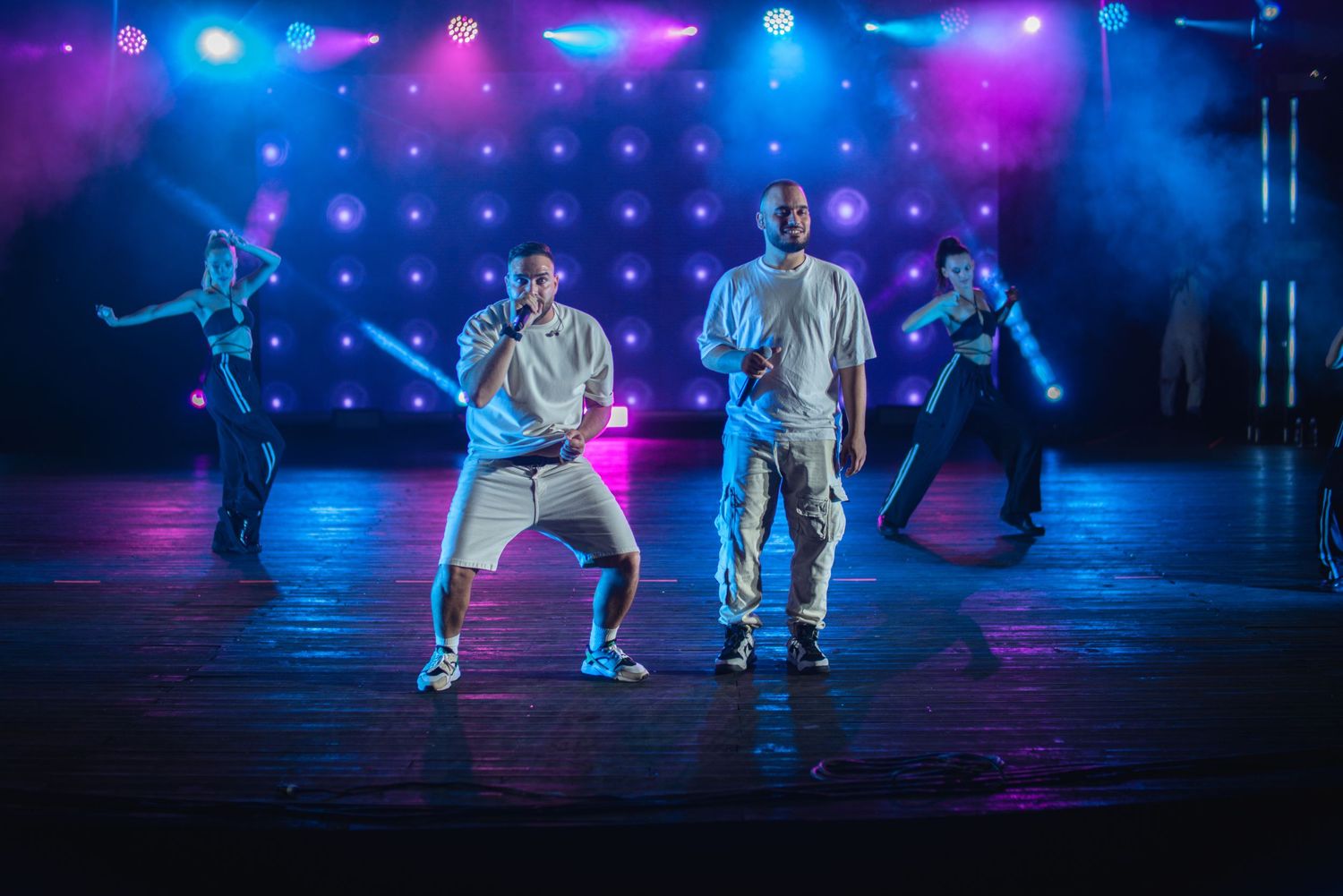 Two performers dancing on stage with backup dancers under vibrant purple and blue LED lights.