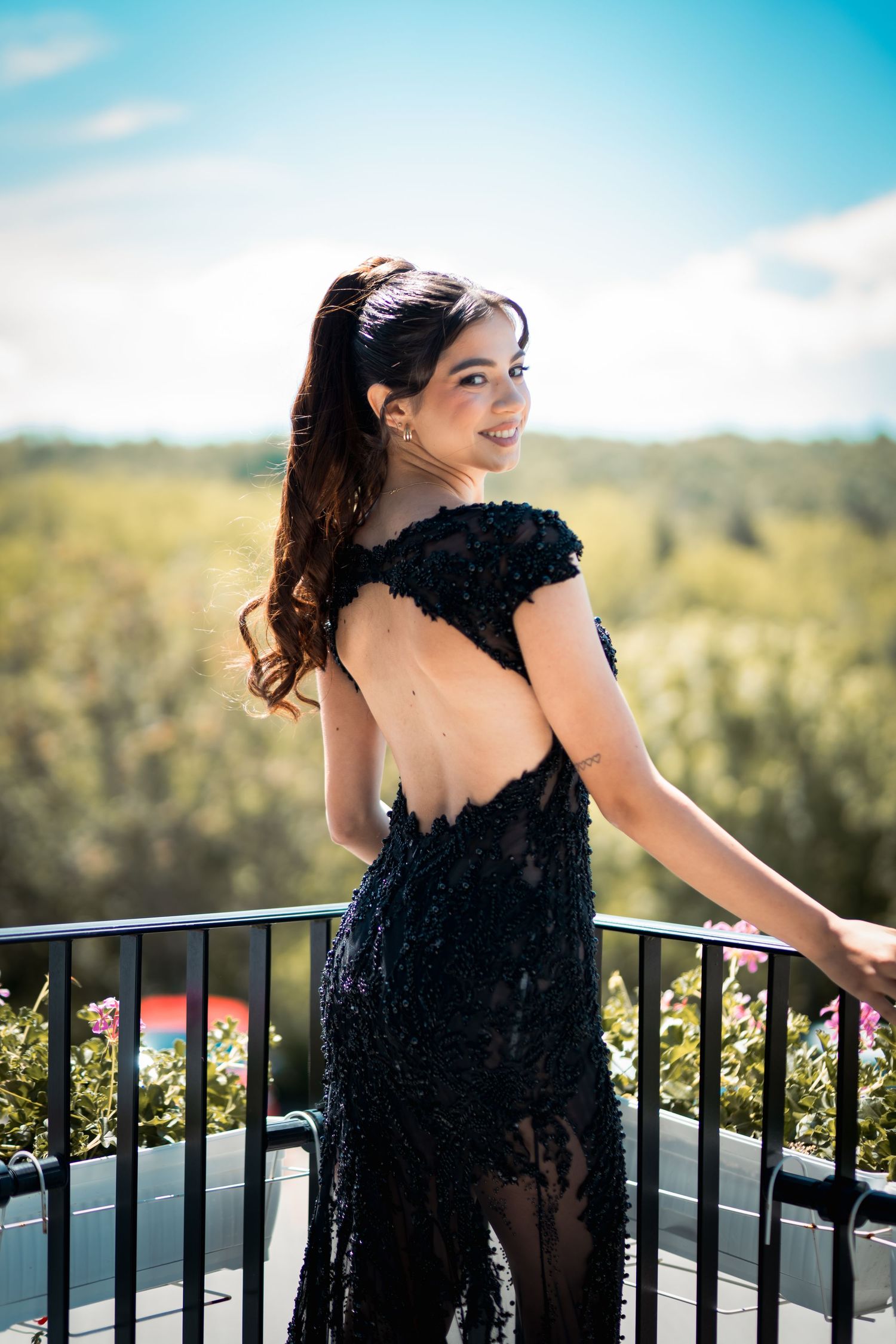 Woman in backless black sequin gown smiling over shoulder on outdoor balcony with scenic view.