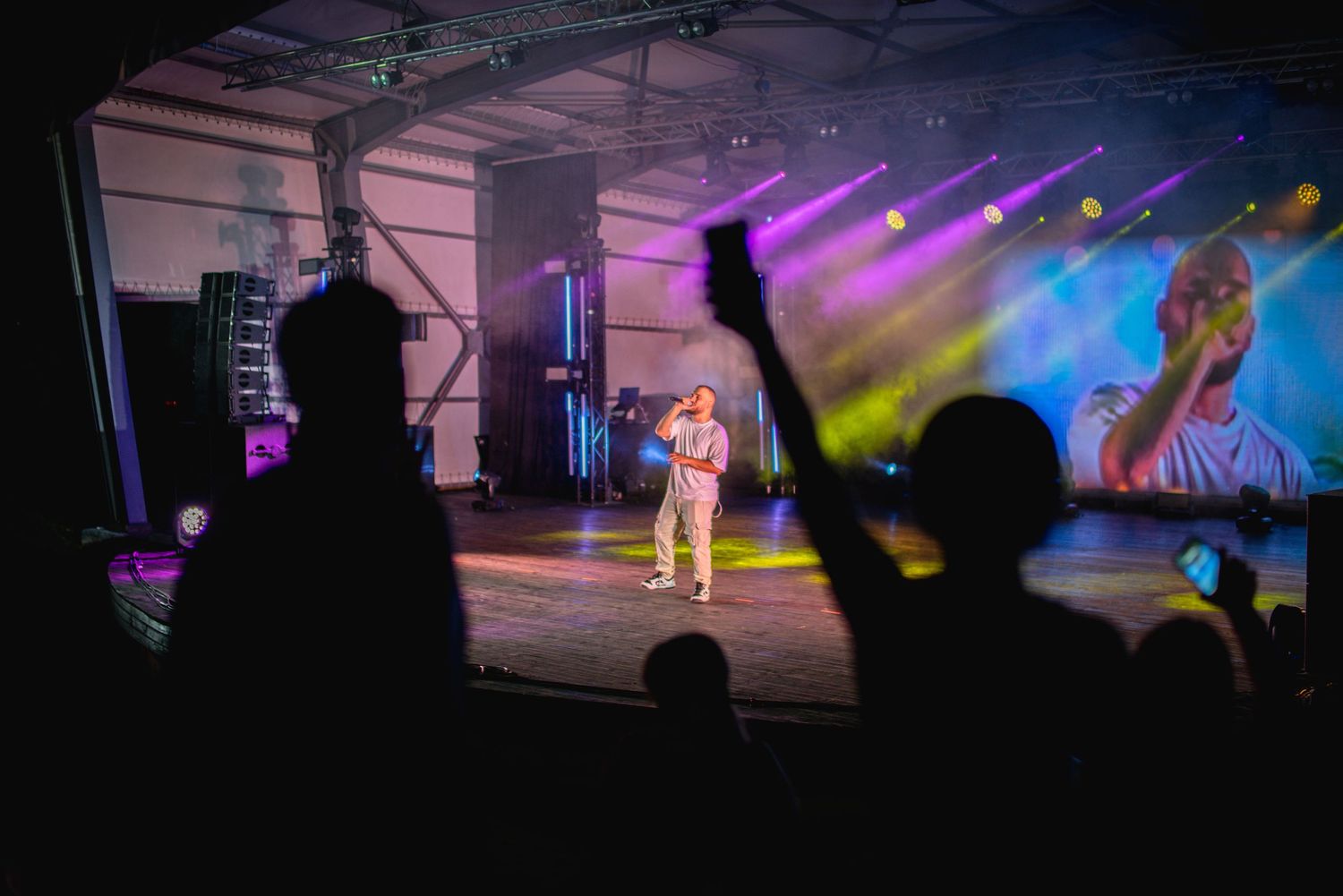 Singer performing on outdoor stage with crowd silhouettes and large video screen projection behind.