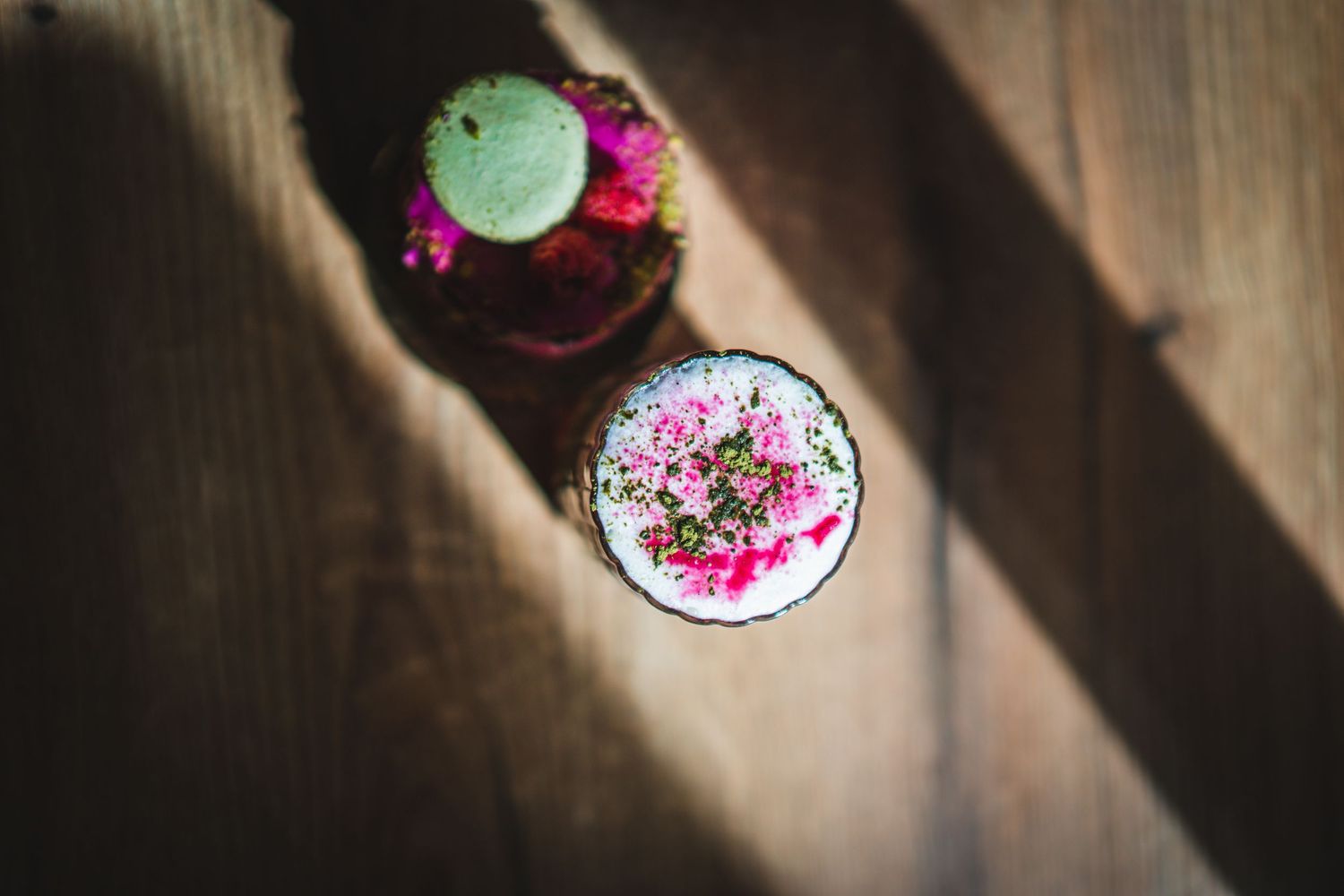 Top view of pink floral foam-topped matcha drink beside dragon fruit dessert on dark wooden surface.