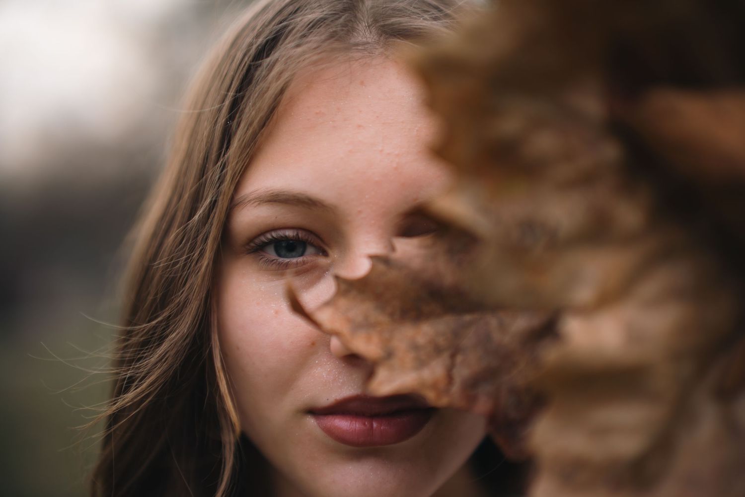 Close-up portrait of young woman with brown hair partially obscured by a dry autumn leaf.