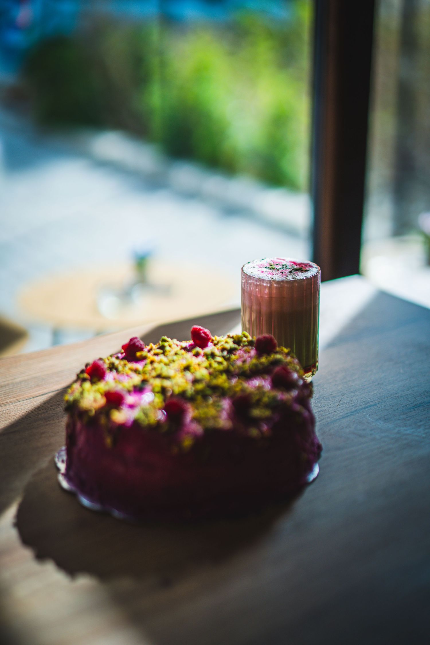 Raspberry pistachio cake topped with fresh berries beside a layered smoothie drink on wooden table.
