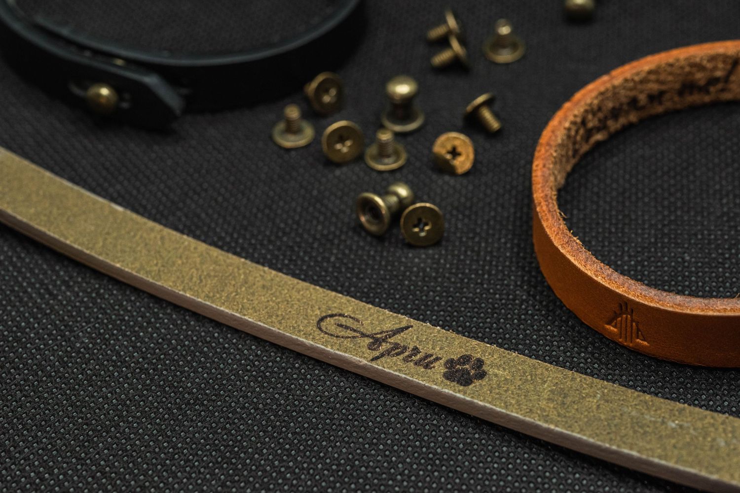 Handcrafted leather bracelet engraved with name Apri and paw print, alongside brass rivets on dark background.