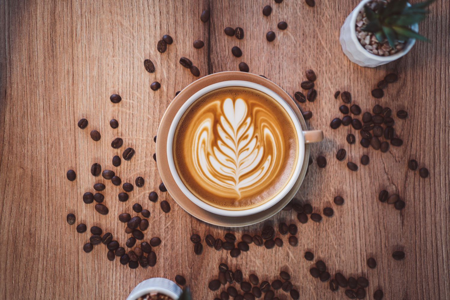 Top view of latte art coffee cup surrounded by scattered roasted coffee beans on wood.
