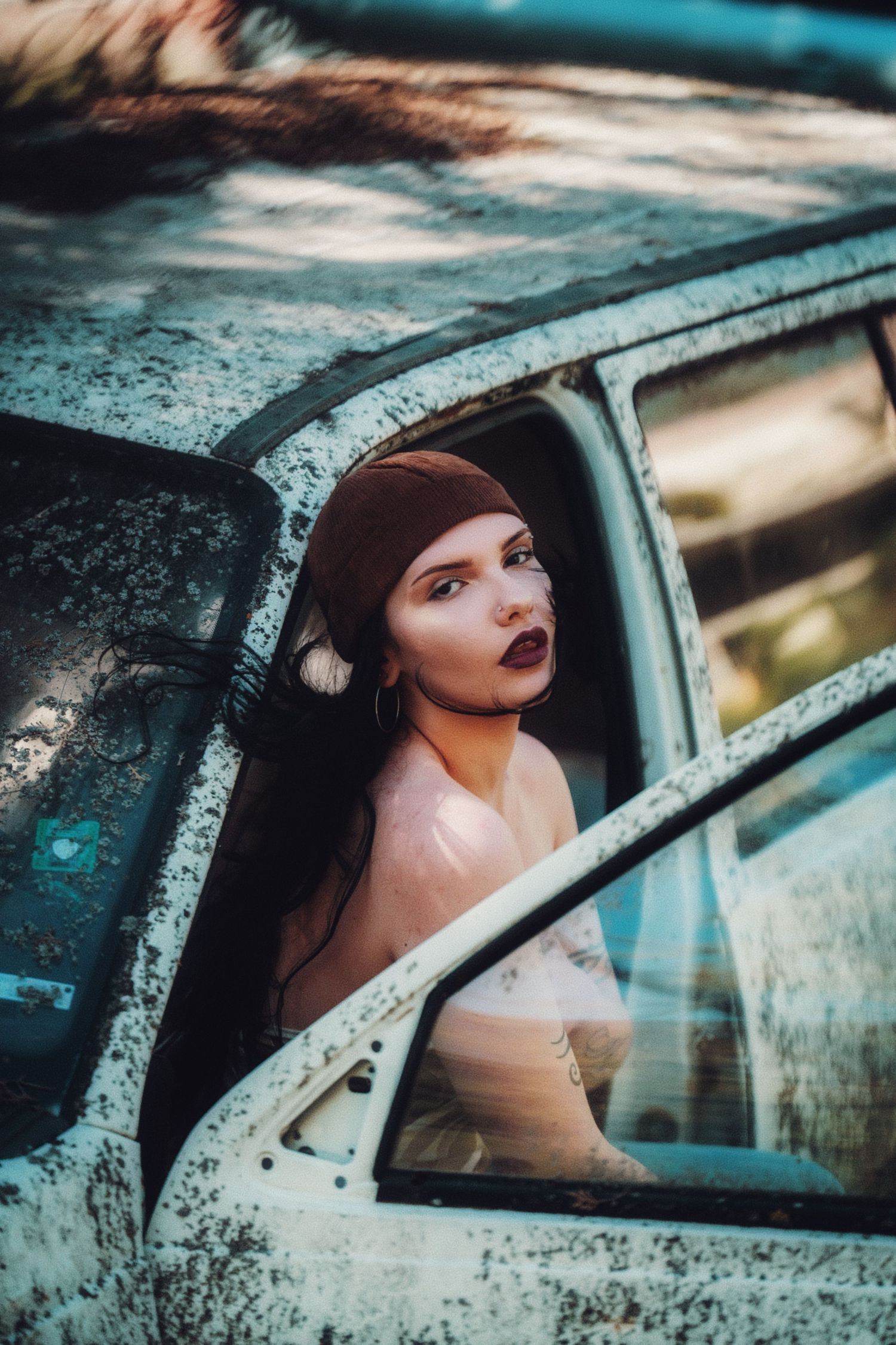 Dark-haired woman in vintage headscarf peers through window of rusty abandoned car with dramatic makeup.