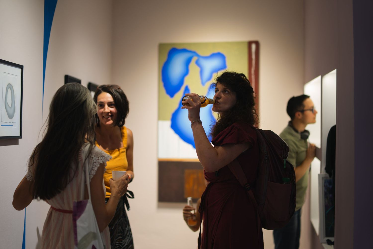 People socializing at an art gallery opening with colorful blue abstract painting on white wall.
