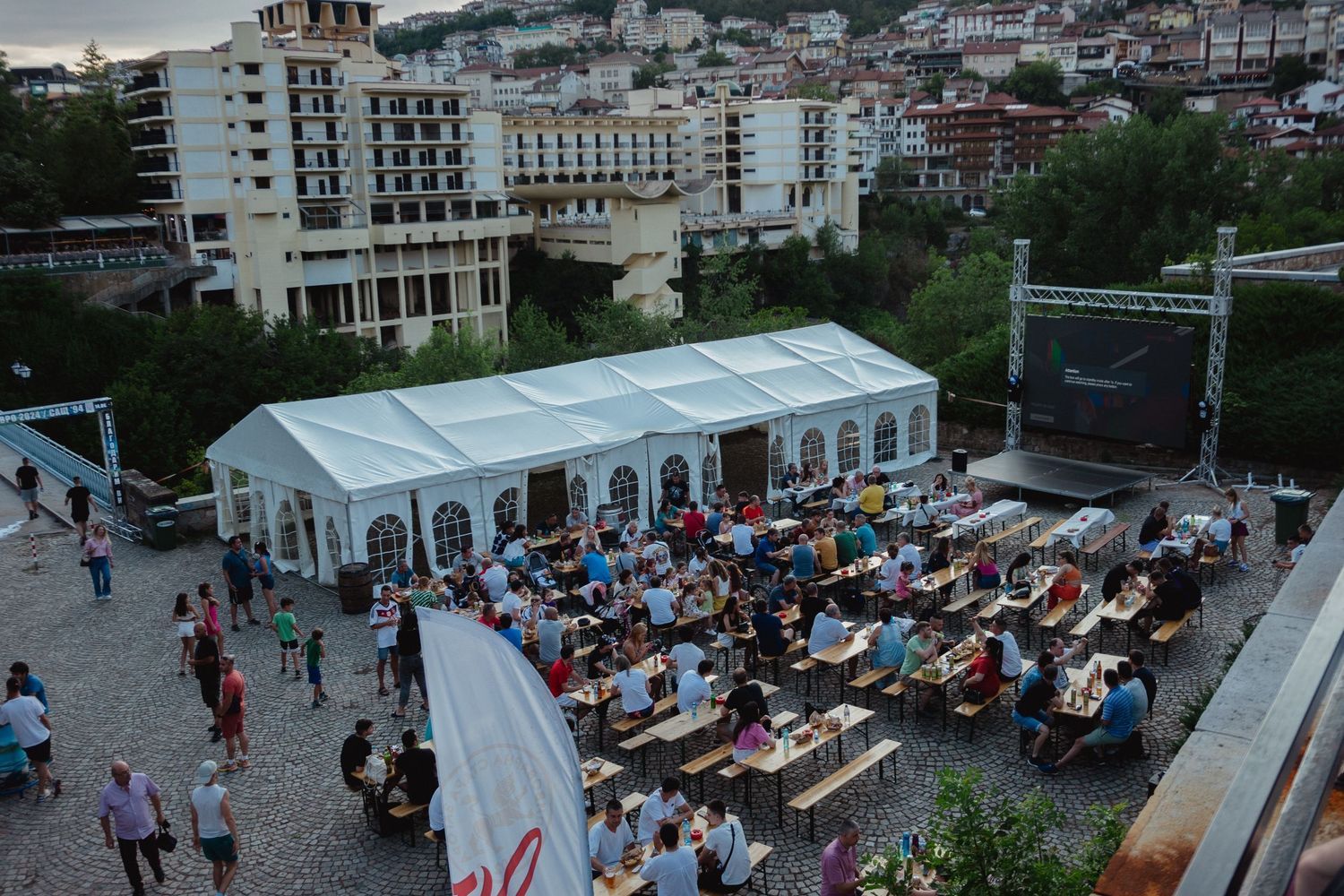 Aerial view of an outdoor festival with crowds seated at long tables under a white tent with a stage and city buildings.